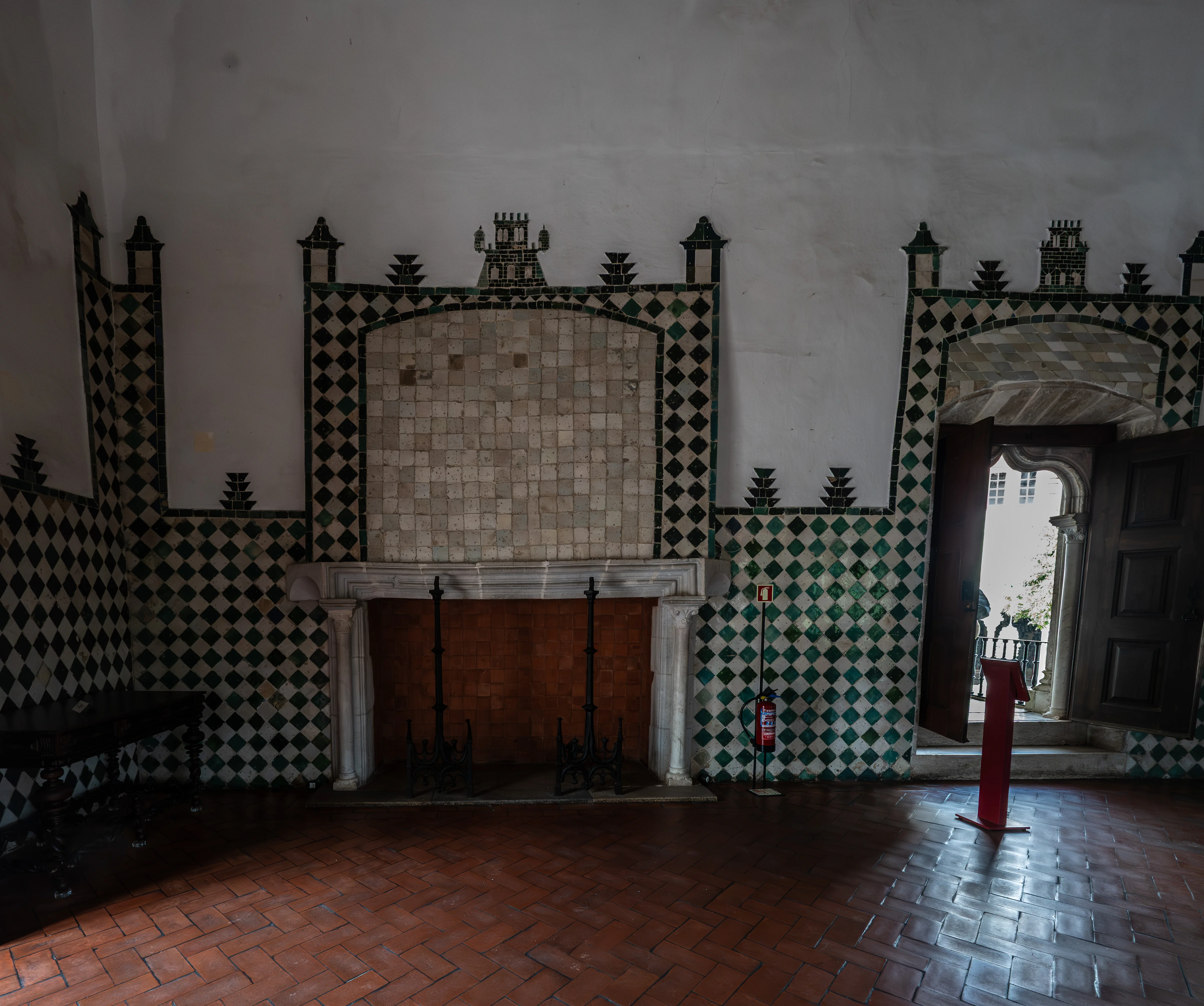 A richly decorated room with a tiled fireplace, diamond-patterned green and white wall tiles, and terracotta flooring, featuring a wooden door on the left and another wooden door open to the outside on the right.