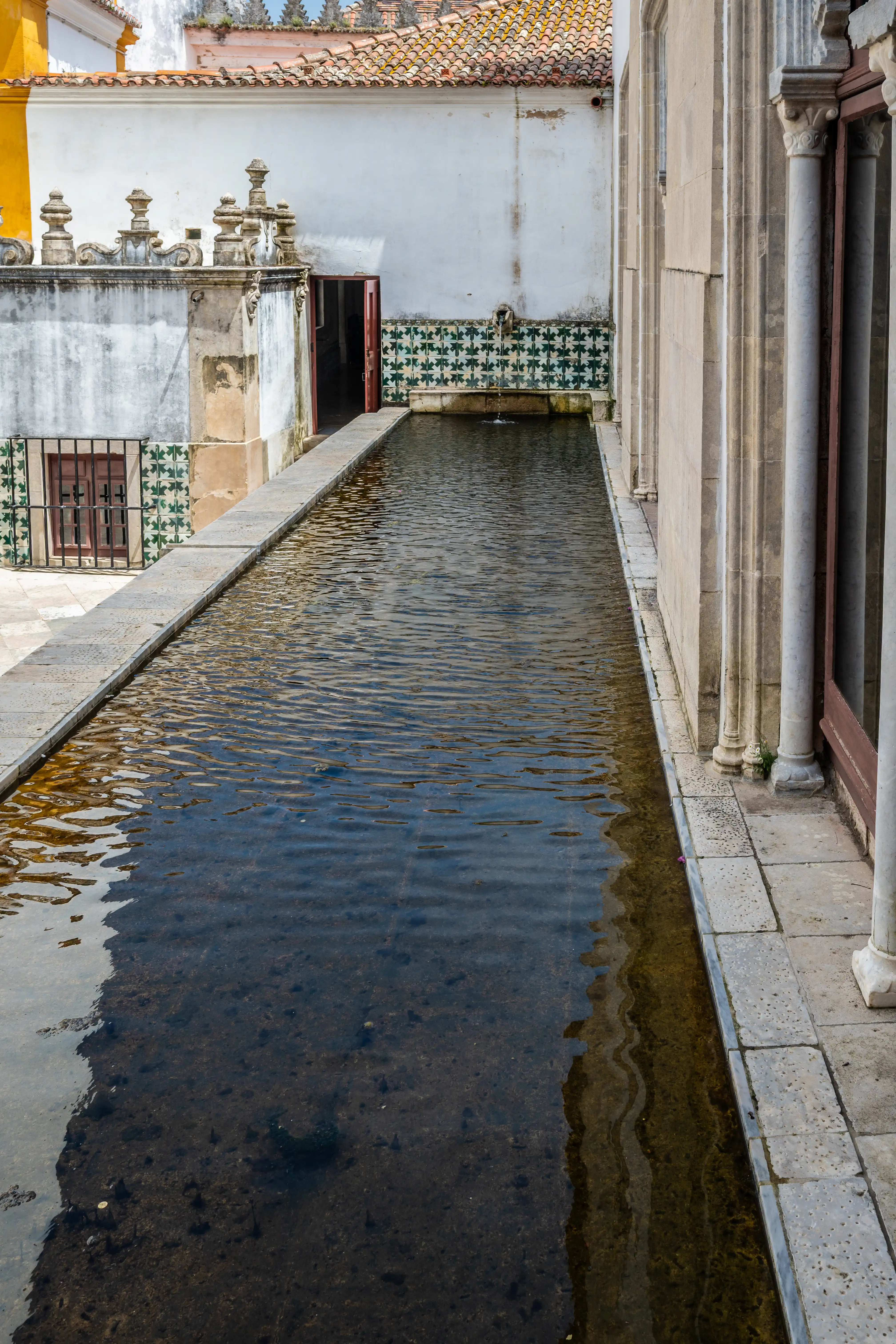 A long, shallow pool of dark water runs alongside a white building featuring patterned green and white azulejo tiles and traditional terracotta roof tiles.