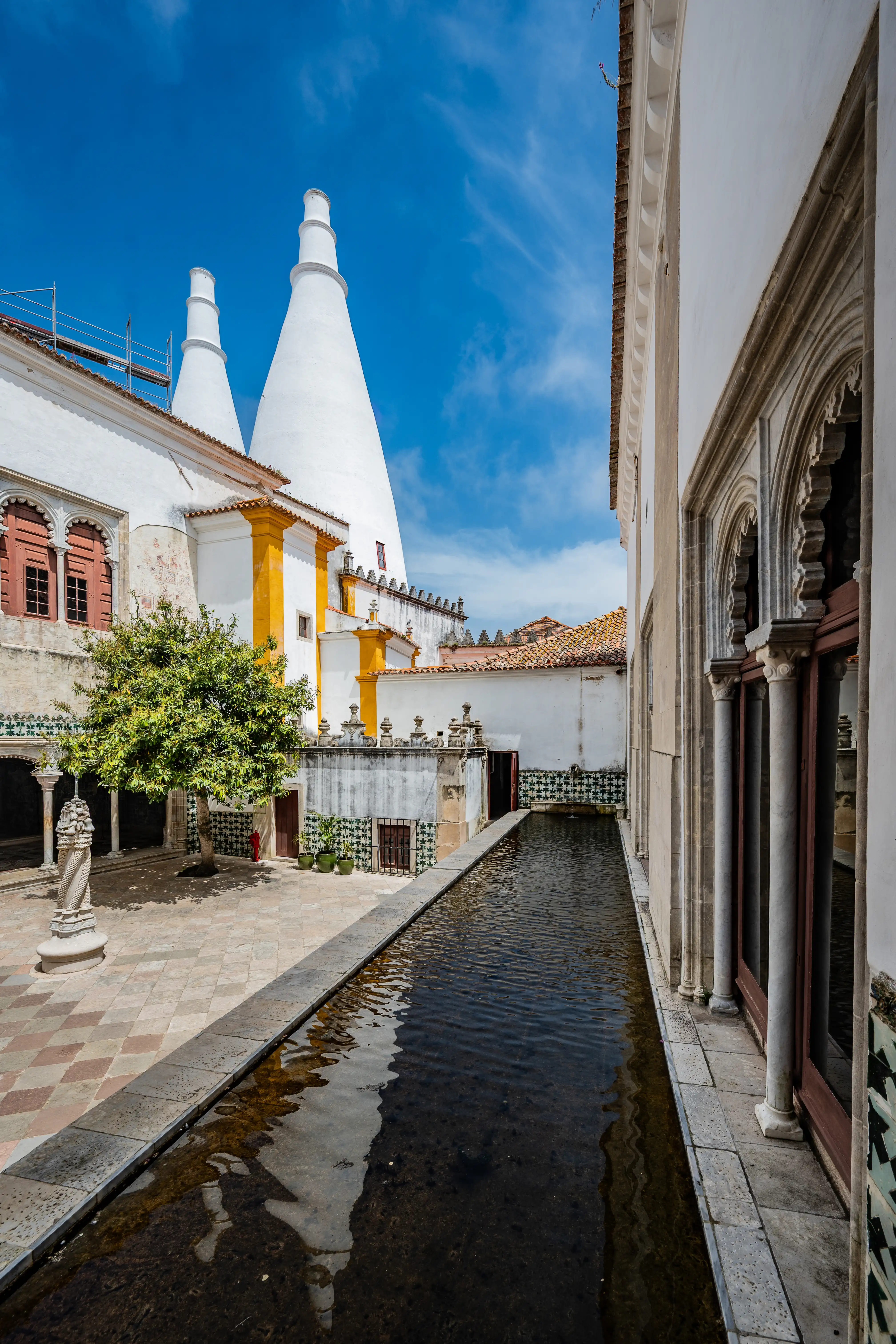 A long, shallow pool of dark water runs alongside a white building featuring patterned green and white azulejo tiles and traditional terracotta roof tiles.