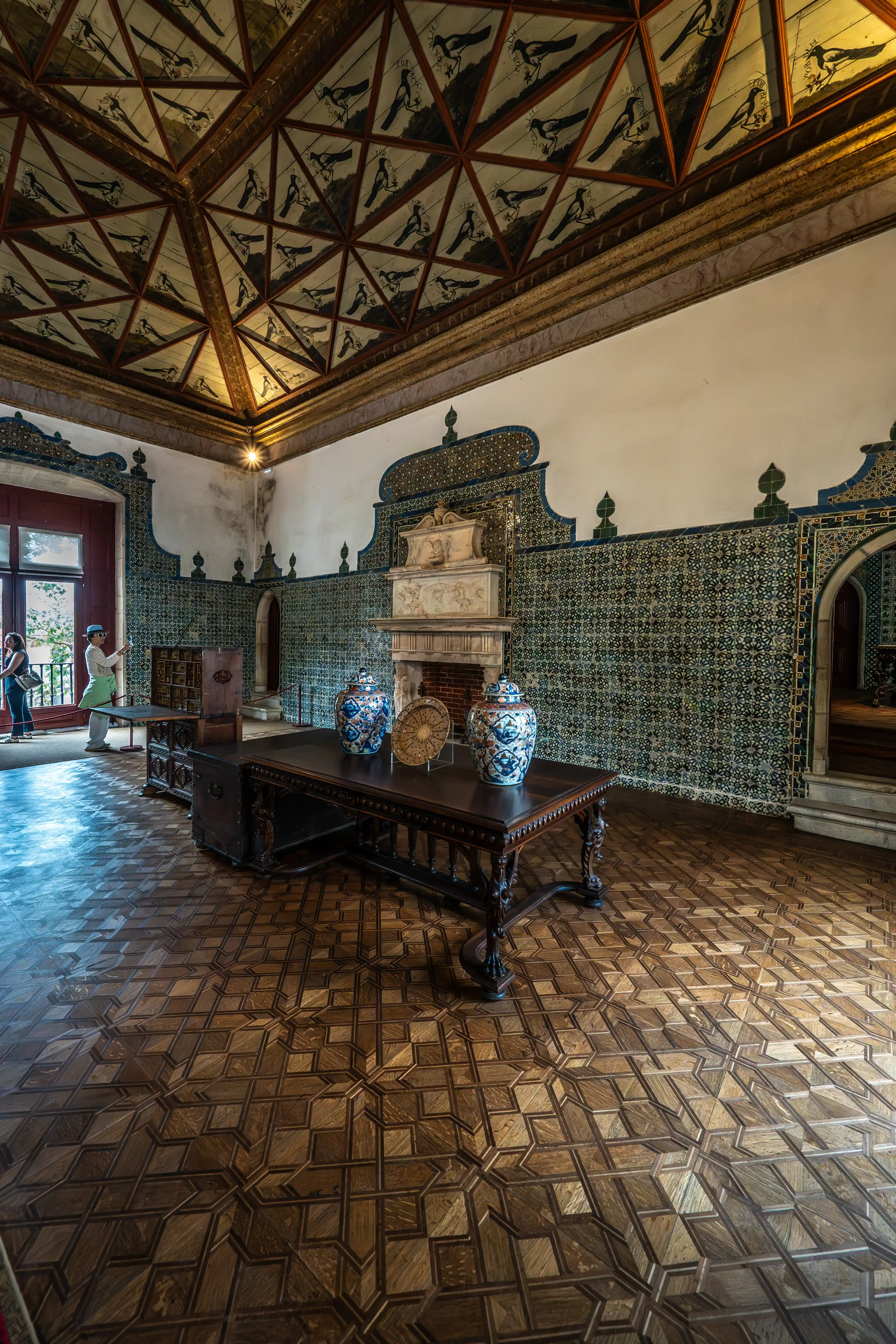 A richly decorated interior room with a bird-themed ceiling, tiled walls, a fireplace, and a wooden table with vases.