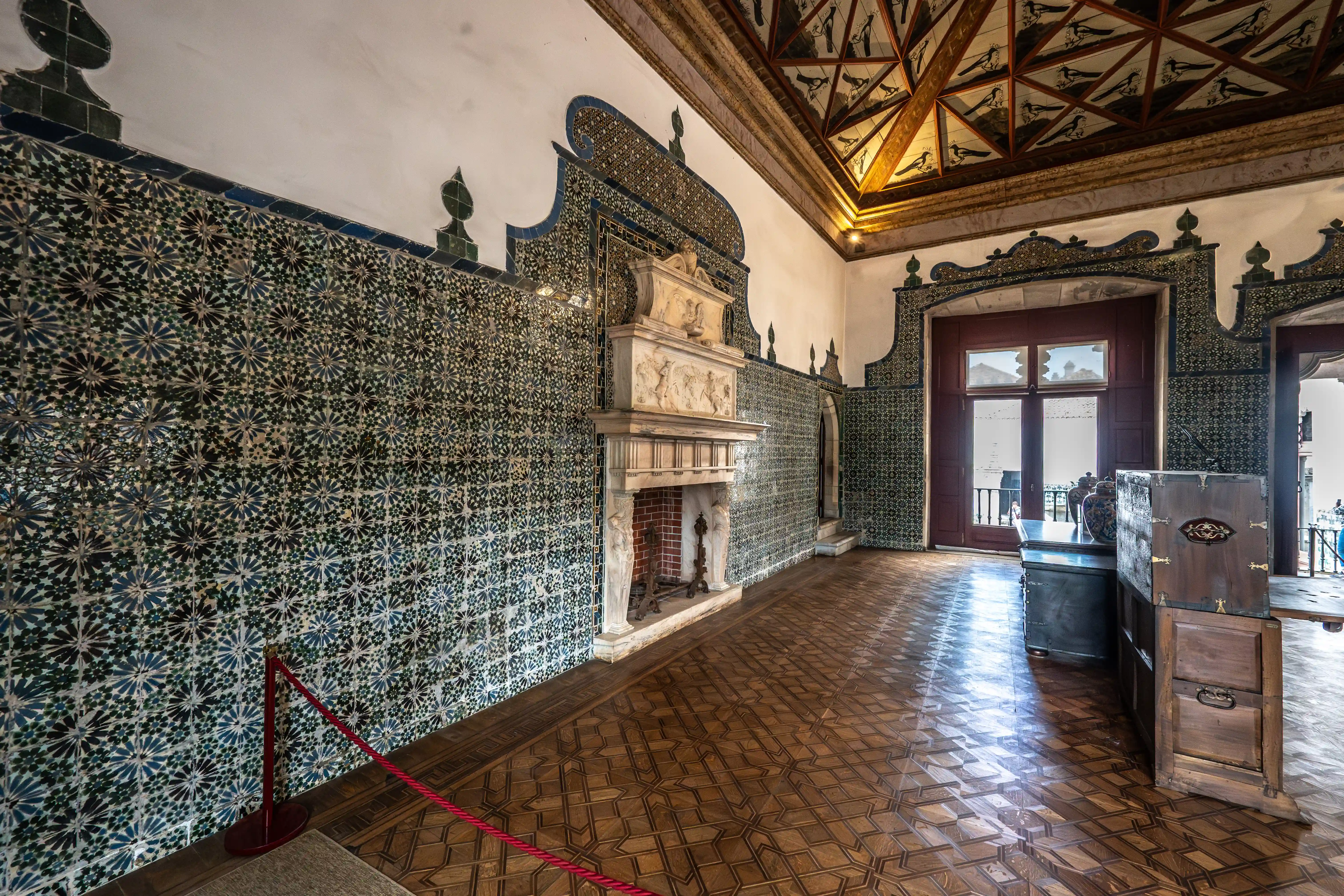 A dimly lit palace room with walls covered in blue and green floral patterned tiles, a decorative stone fireplace, a patterned wooden floor, and a large window with a view of the outside.