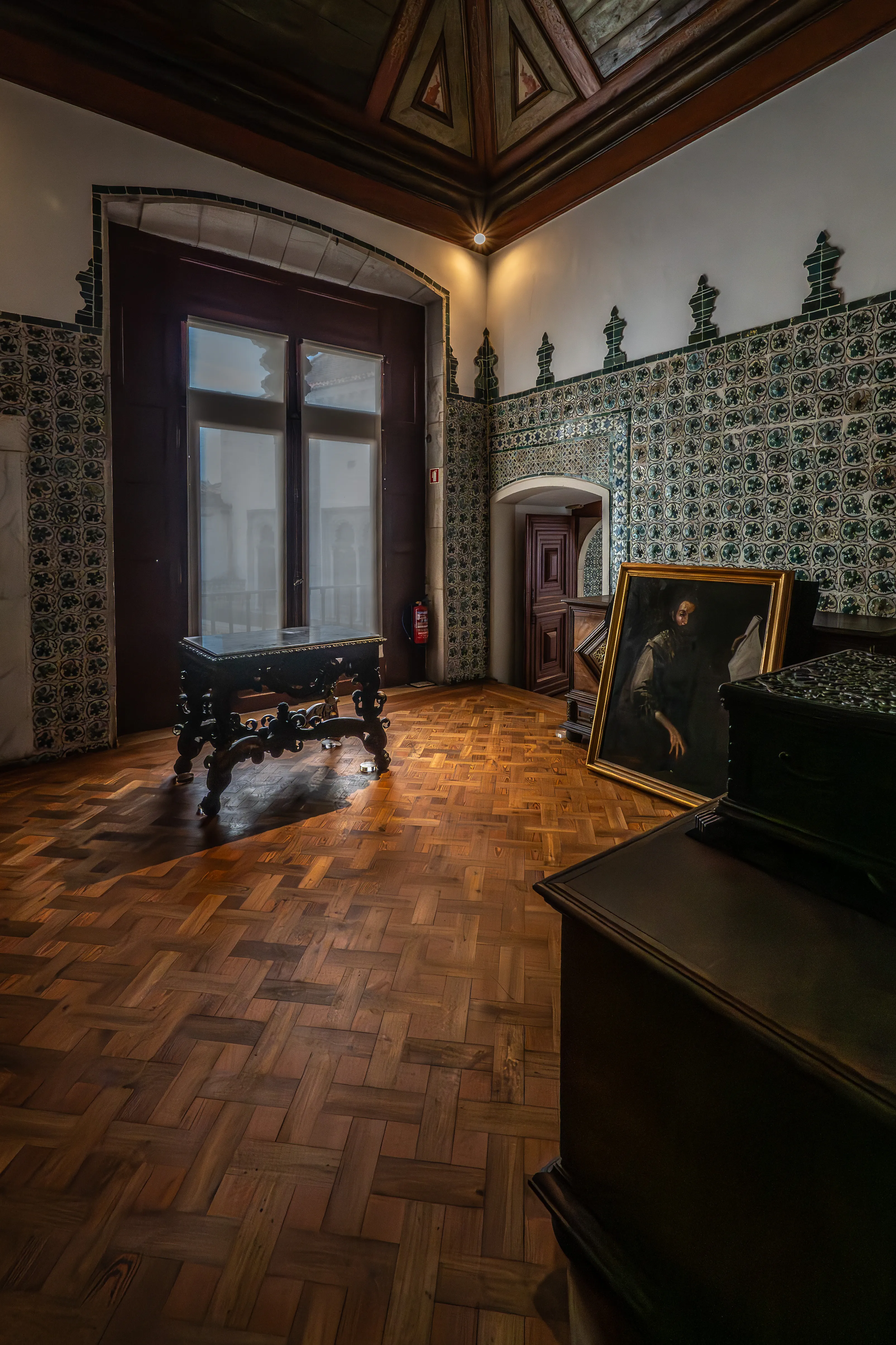 A dark, moody interior of a room featuring a tall, bright, cross-paned window with a decorative table in front of it. Ornate tiled walls and a framed painting are visible, along with a wooden floor and ceiling details.
