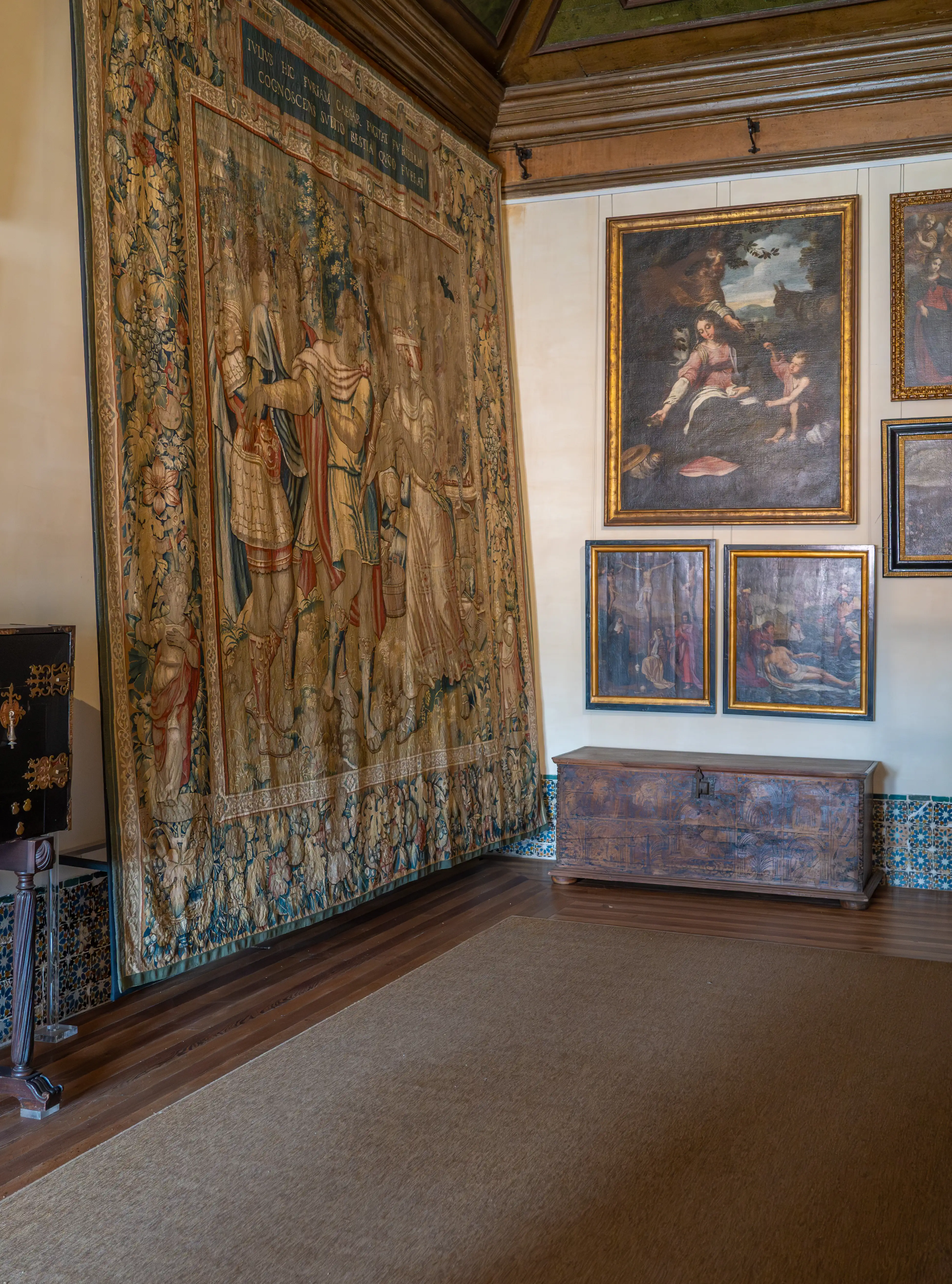 A large, ornately woven tapestry depicting figures hangs on the left wall of a richly decorated room. On the right wall, several framed paintings are displayed above a wooden chest. A brown rug covers a portion of the floor.
