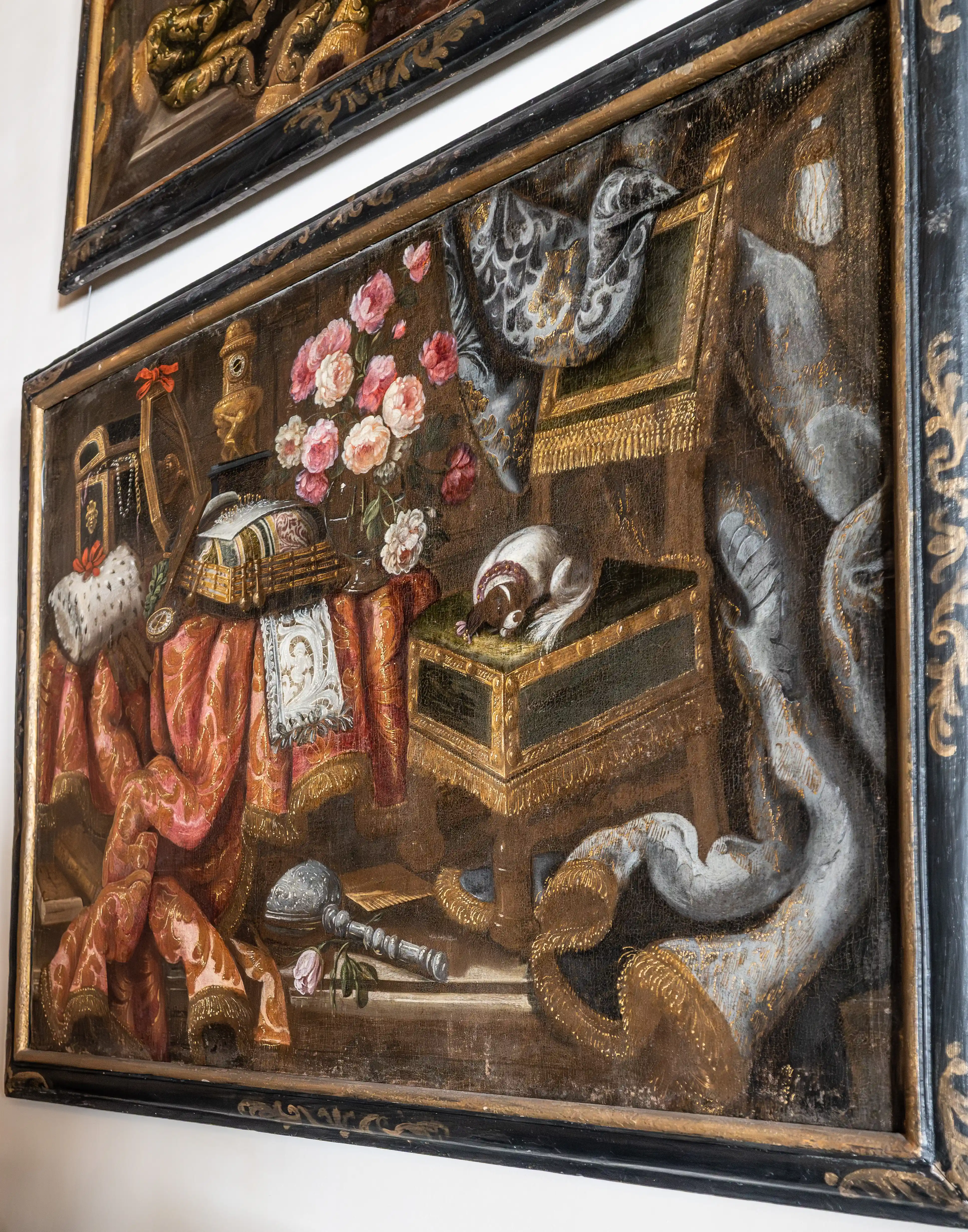 A framed oil painting of a still life arrangement on a table, featuring a small dog sleeping on a green-topped chest. To the left, opulent red and gold fabric is draped, with a fur-trimmed hat and jewellery visible. A vase of pink and white roses sits in the centre, alongside a mirror and other decorative items. To the right, a large, draped grey fabric cascades down, with a golden fringe.