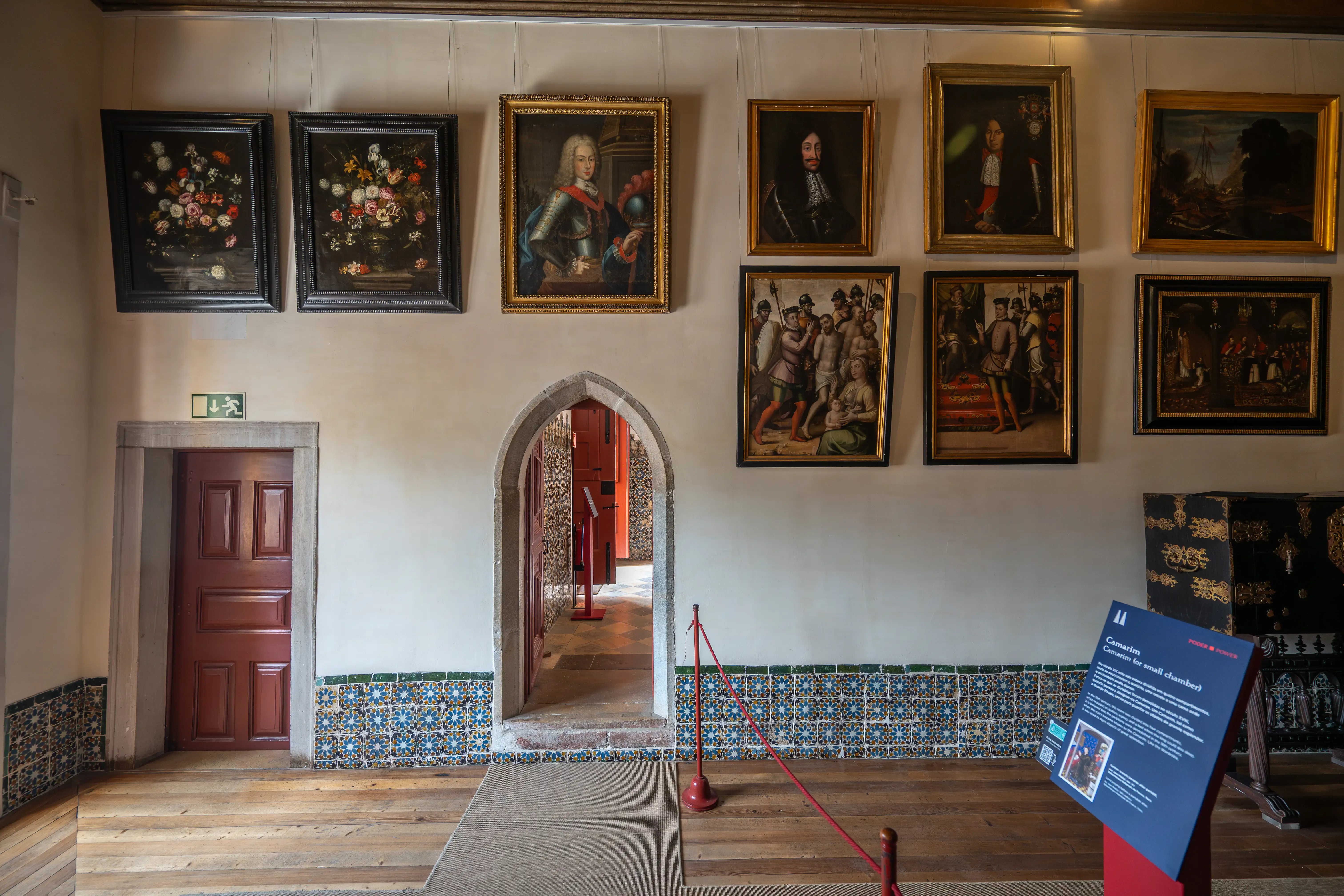 Interior view of a historic hallway with framed paintings on the wall, decorative tiles, wooden floor, an arched doorway, and an information board.