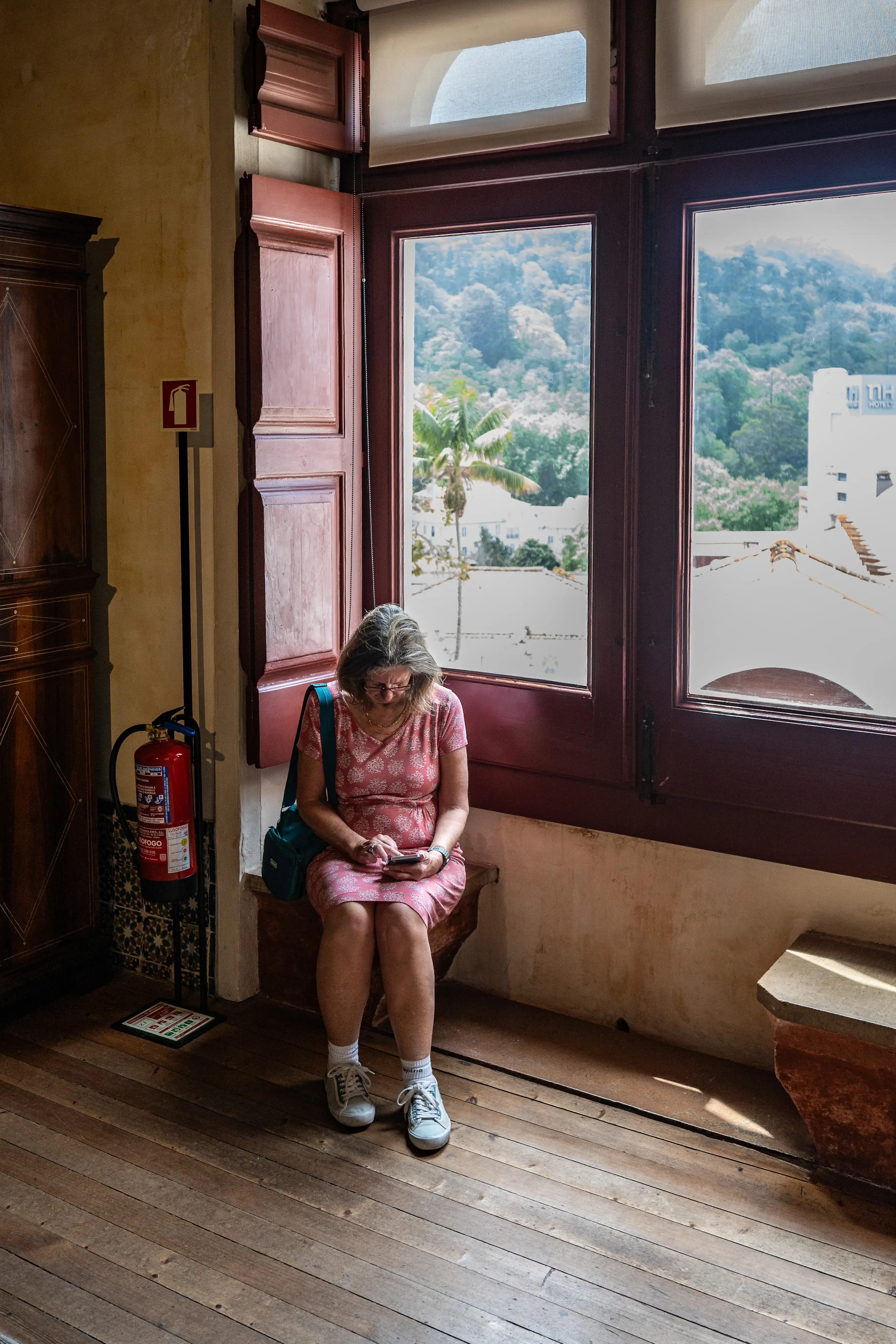A woman wearing glasses and a pink dress sits by a large window, engrossed in her mobile phone.