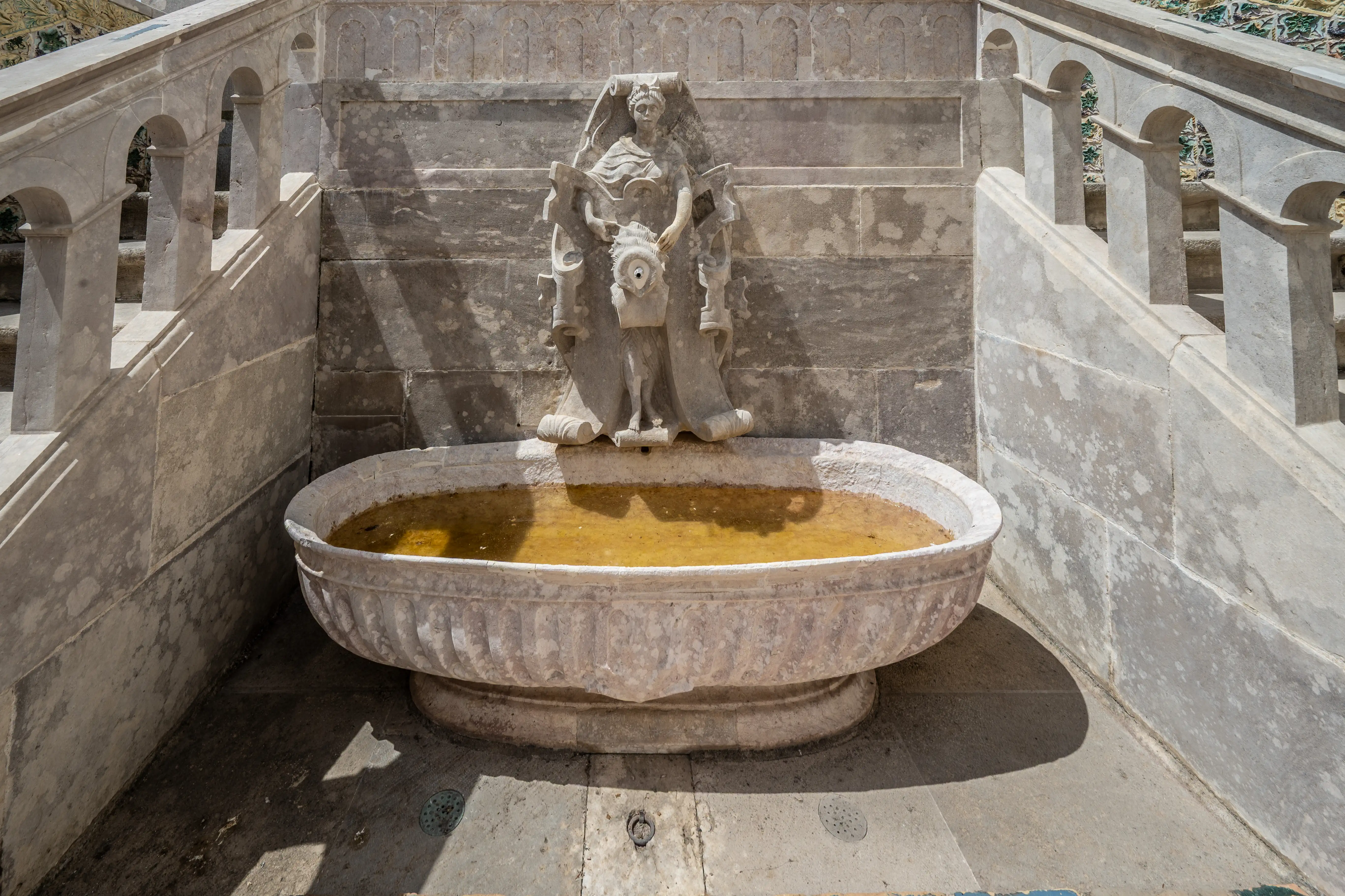 A close-up view of an ancient stone fountain with a carved human-like figure, situated at the base of a grand stone staircase. The fountain basin holds murky yellow water. The staircase features ornate balustrades with arches and decorative tiled sections on the walls.
