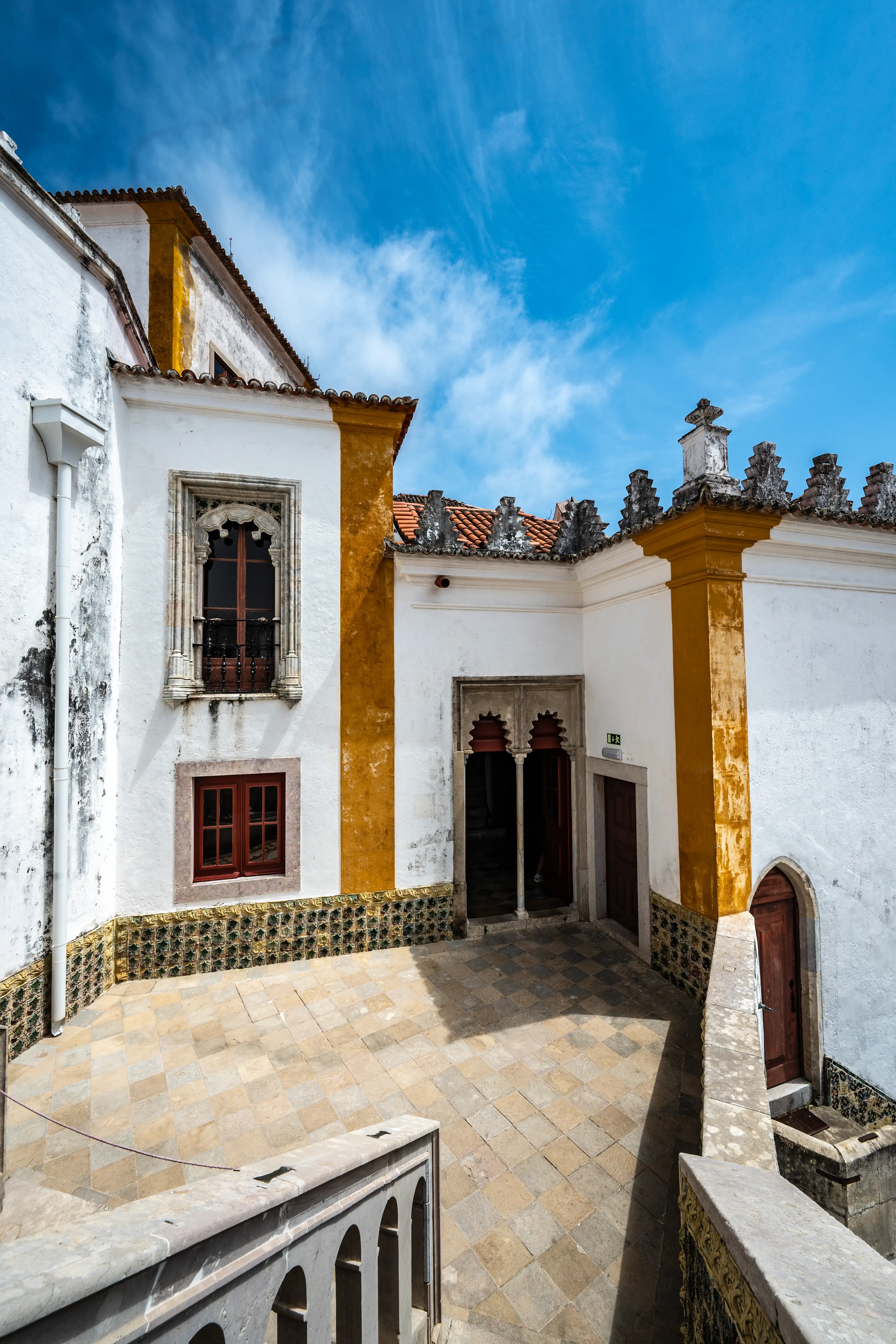 A view from above into a courtyard at Pena Palace, Sintra. White palace walls with yellow accents and red-trimmed windows are on the left. People walk on a stone path towards an archway in the middle ground, with a green, tree-covered hill topped by the Moorish Castle in the background under a blue sky.