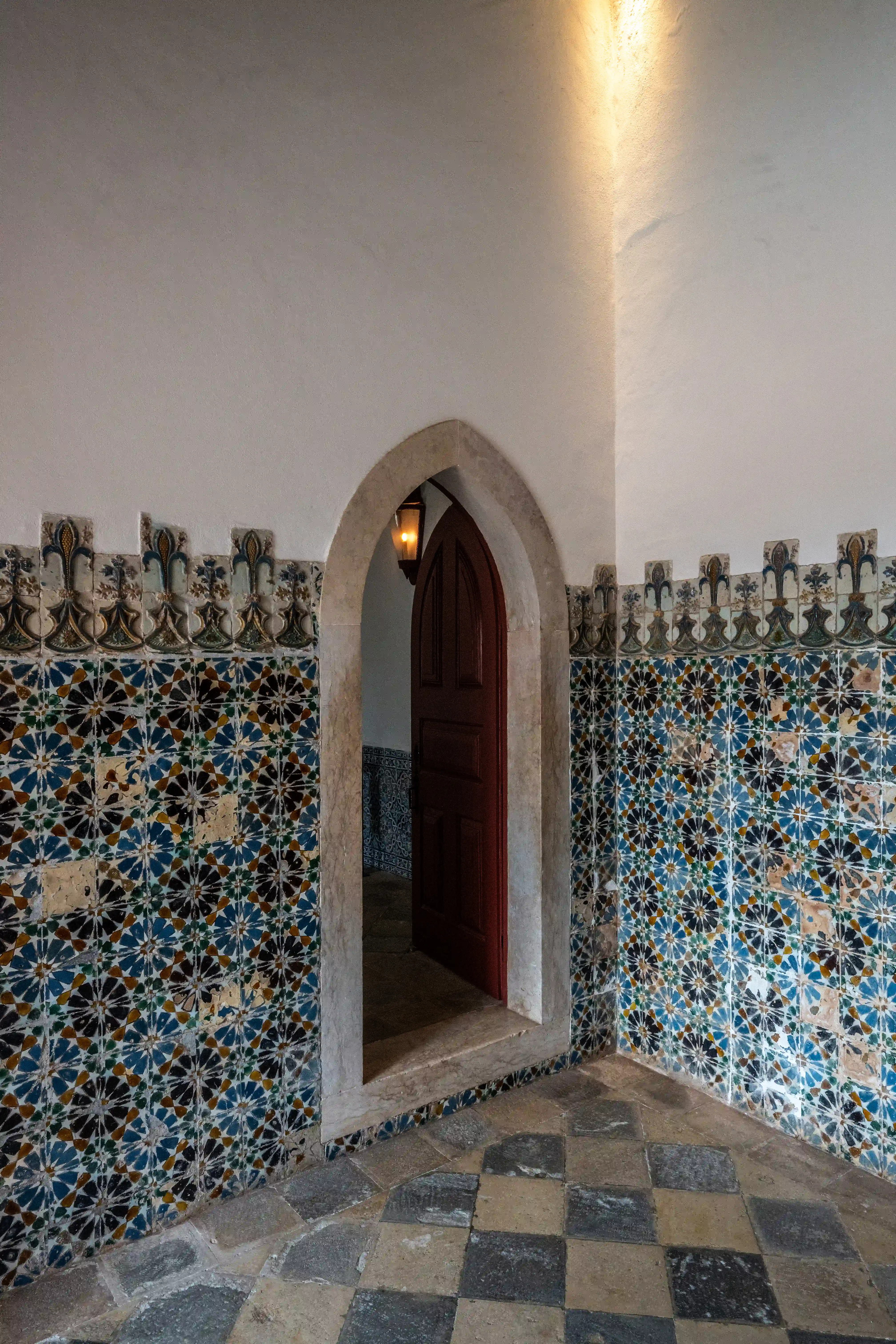 Interior of a historic corridor with ornate patterned tiles and an arched doorway leading to a dark wooden door.