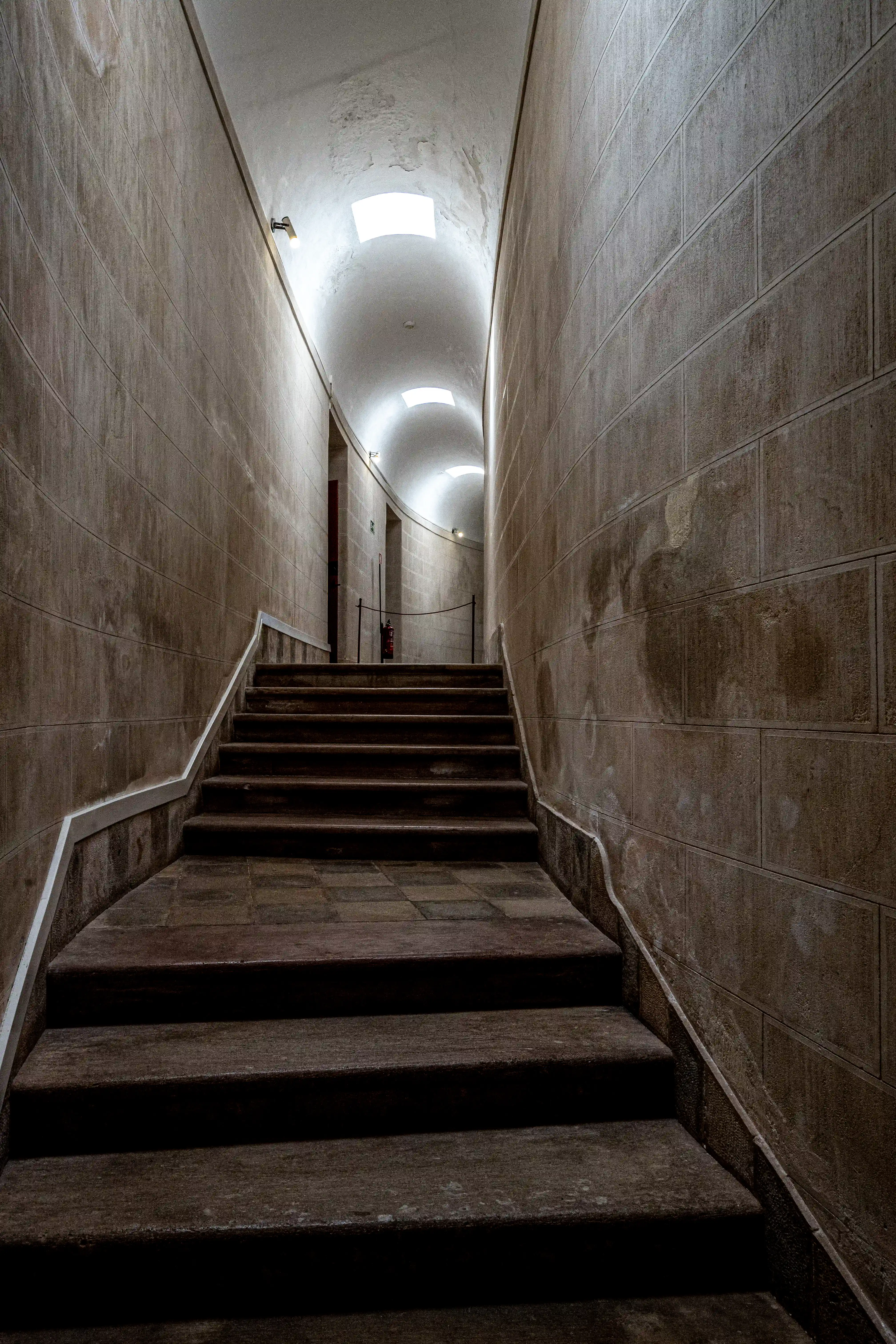 An indoor view of a curved stone staircase leading upwards into a vaulted corridor with skylights.