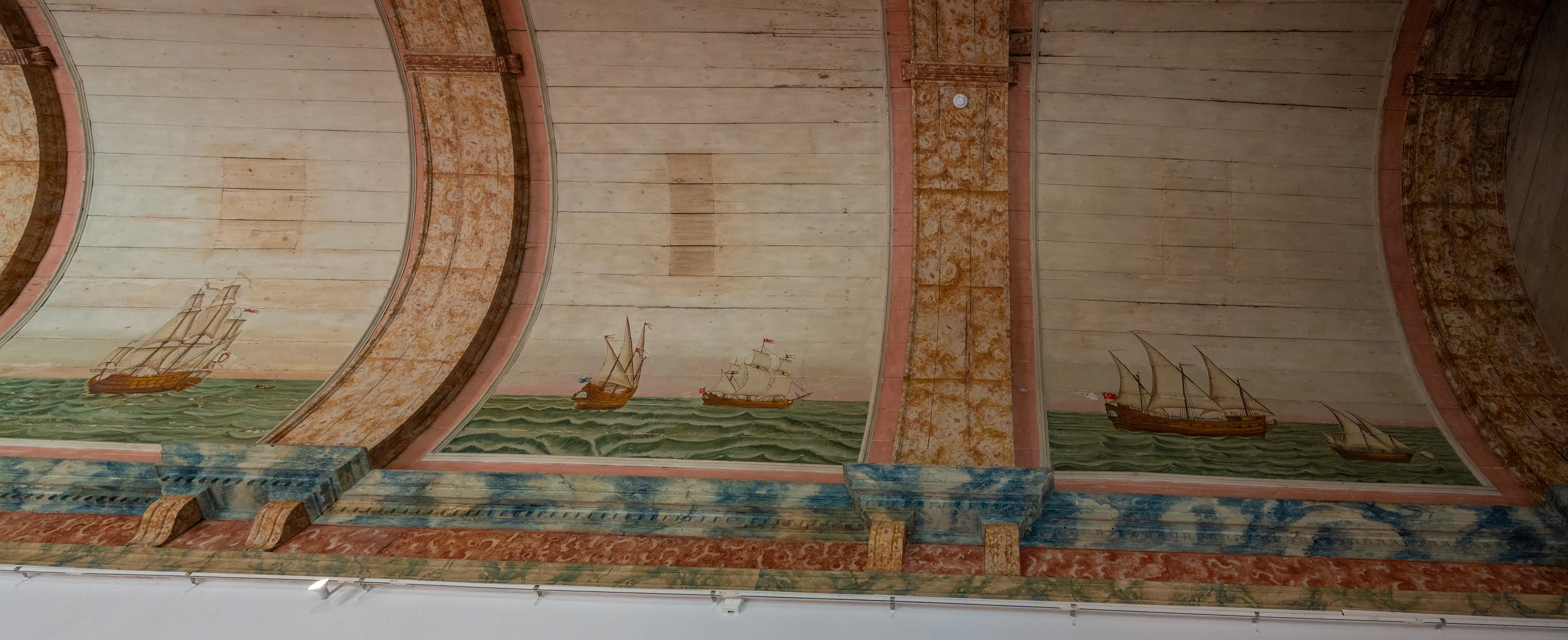 Painted wooden ceiling with arched sections, each showing a different sailing ship on a green sea. Decorative bands of red, blue, and patterned orange frame the maritime scene.