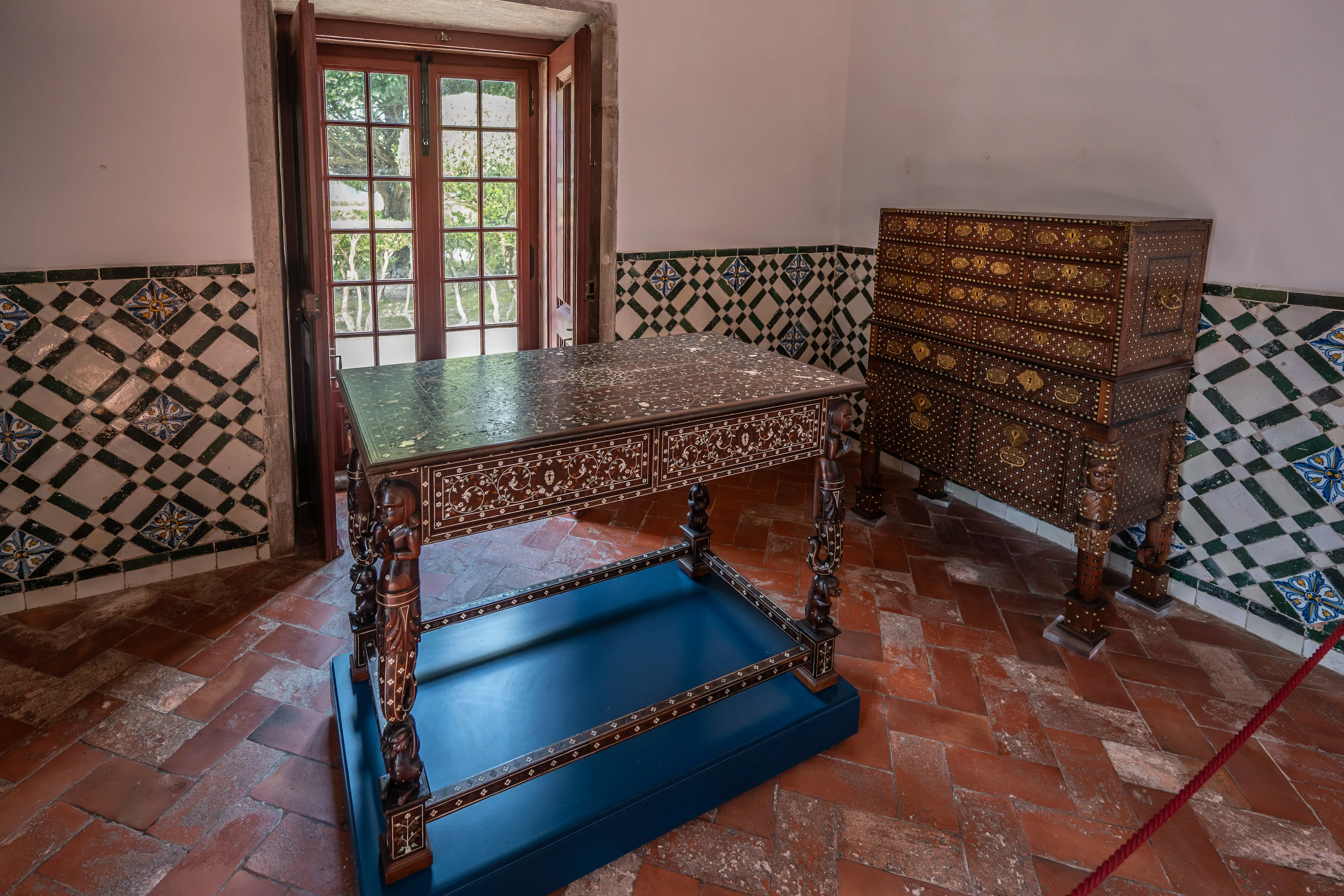 A room featuring an antique inlaid wooden table with carved legs on a blue plinth, a decorative chest of drawers, and walls adorned with geometric tiles. French doors with multiple panes are visible on the left side, and the floor is tiled with terracotta in a herringbone pattern.