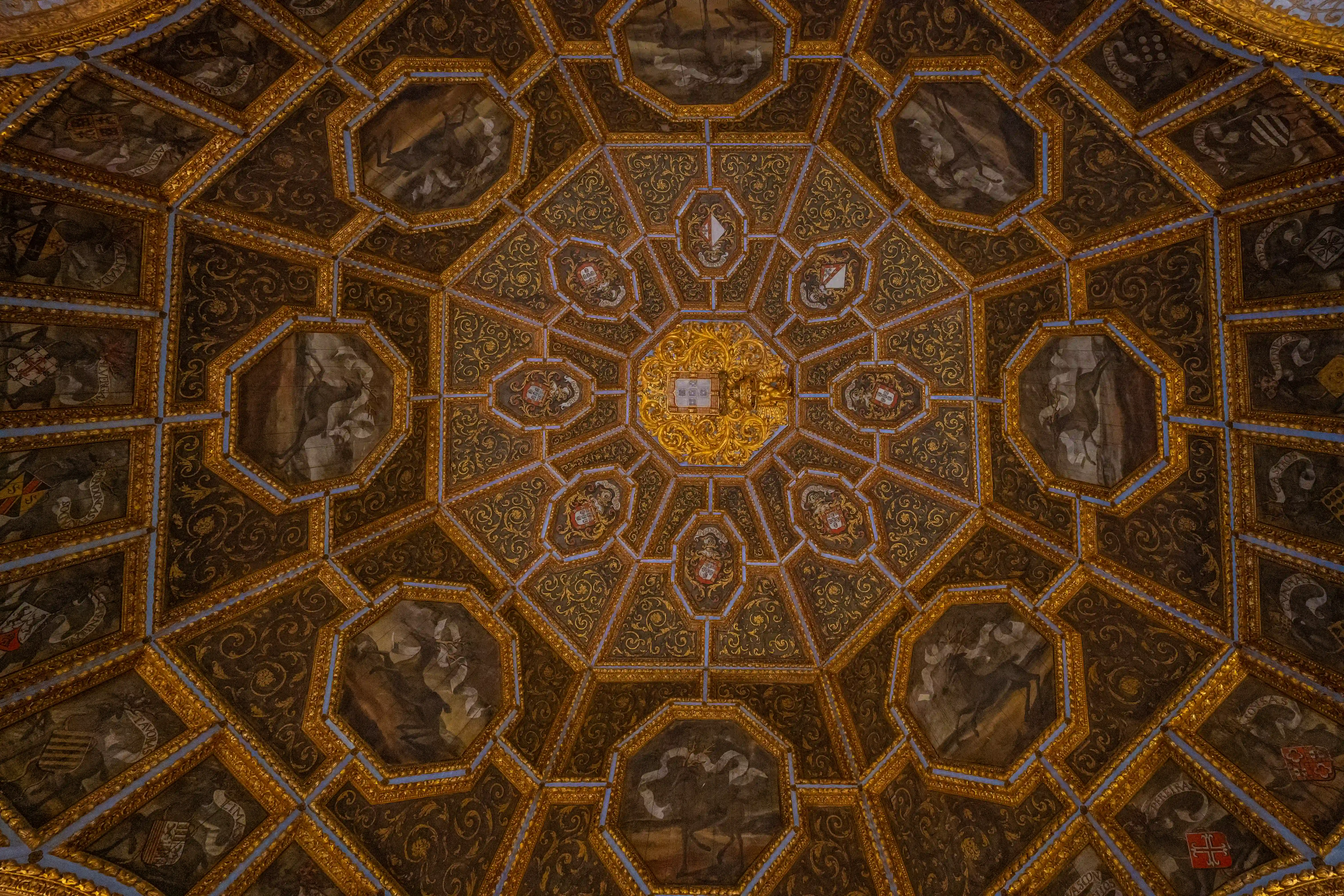 An elaborate octagonal ceiling with dark and gold geometric patterns, featuring painted scenes and heraldic shields within octagonal and square panels.