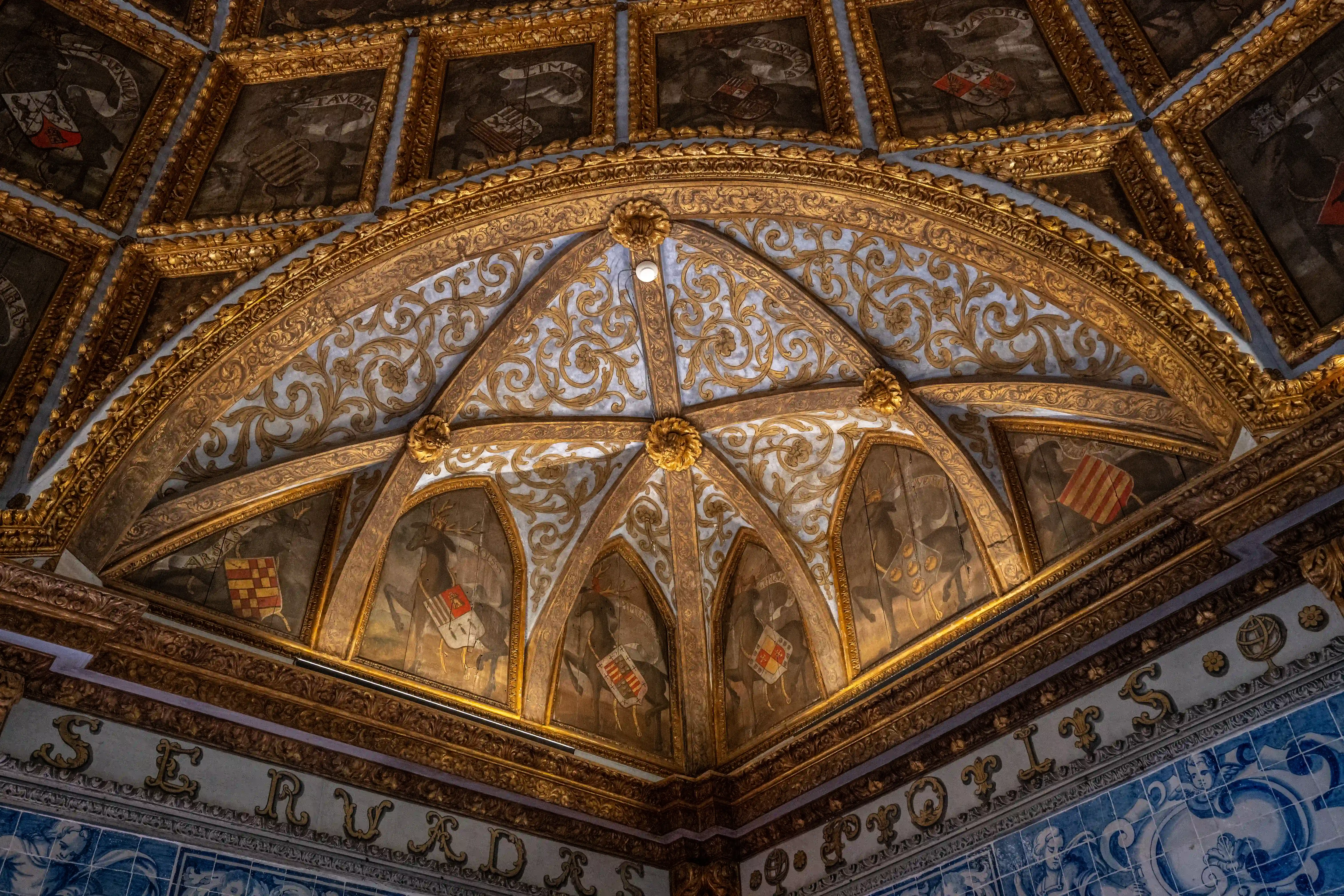 Low-angle view of a richly decorated vaulted ceiling. Golden mouldings frame panels containing paintings of heraldic crests, shields, and figures, interspersed with scrolling gold patterns on a light blue background. A border of blue and white azulejo tiles with an inscription runs along the bottom of the image.
