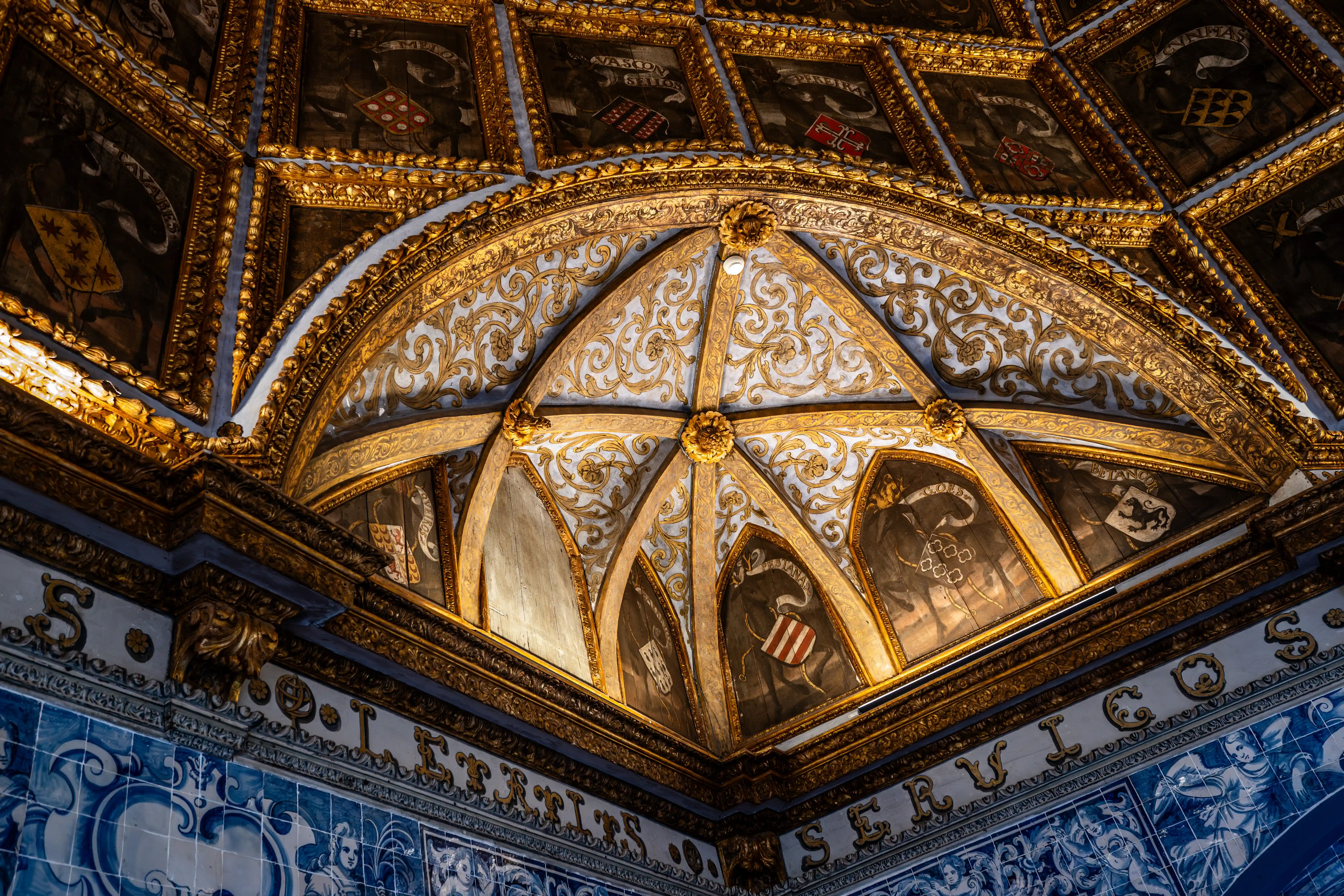 A close-up view of a highly decorated ceiling featuring a golden ribbed vault with blue panels, ornate gold scrollwork, and framed shields displaying coats of arms. A decorative frieze with gilded lettering sits above blue and white patterned tiles on the wall below.