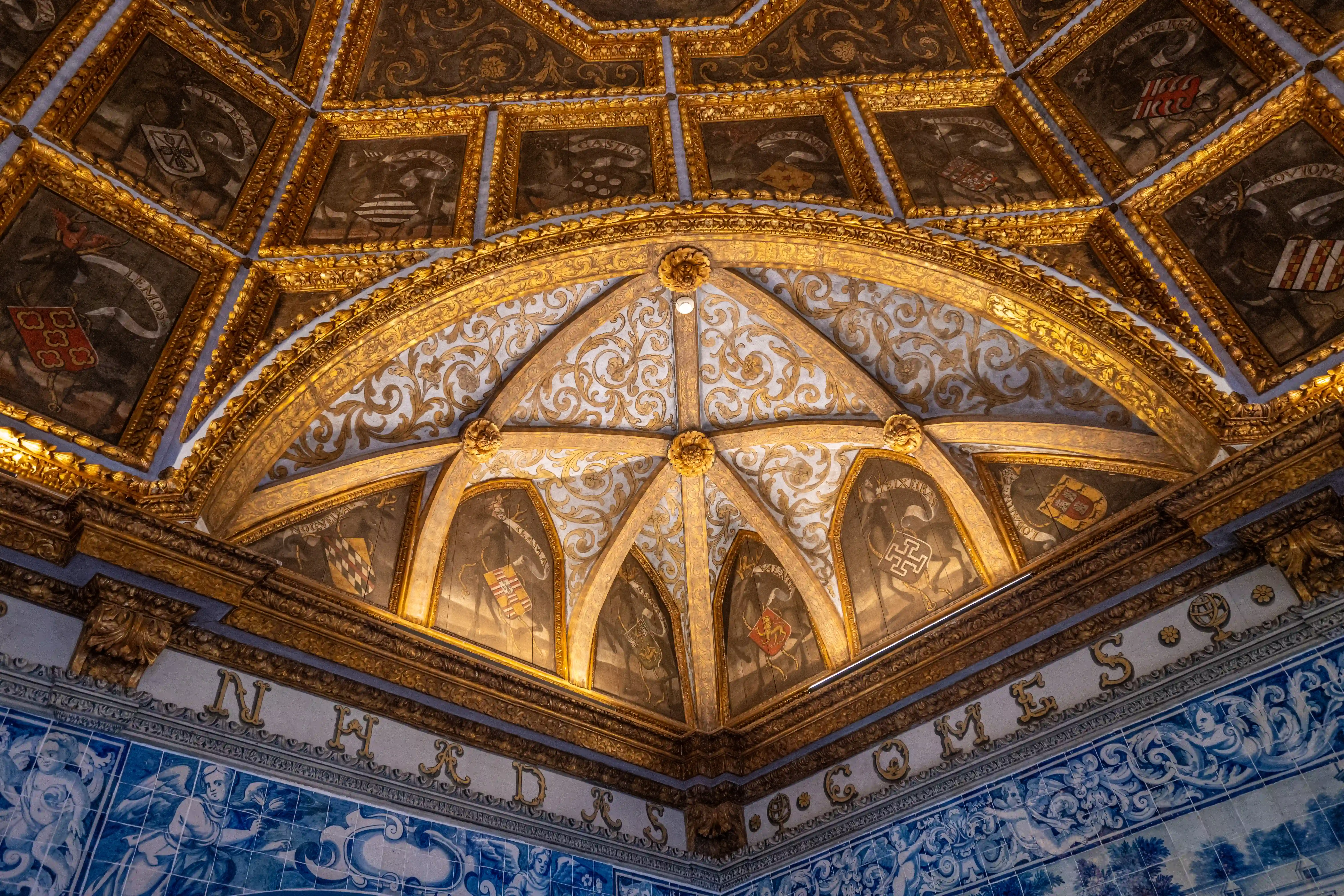 A close-up view of a highly decorated ceiling featuring a golden ribbed vault with blue panels, ornate gold scrollwork, and framed shields displaying coats of arms. A decorative frieze with gilded lettering sits above blue and white patterned tiles on the wall below.