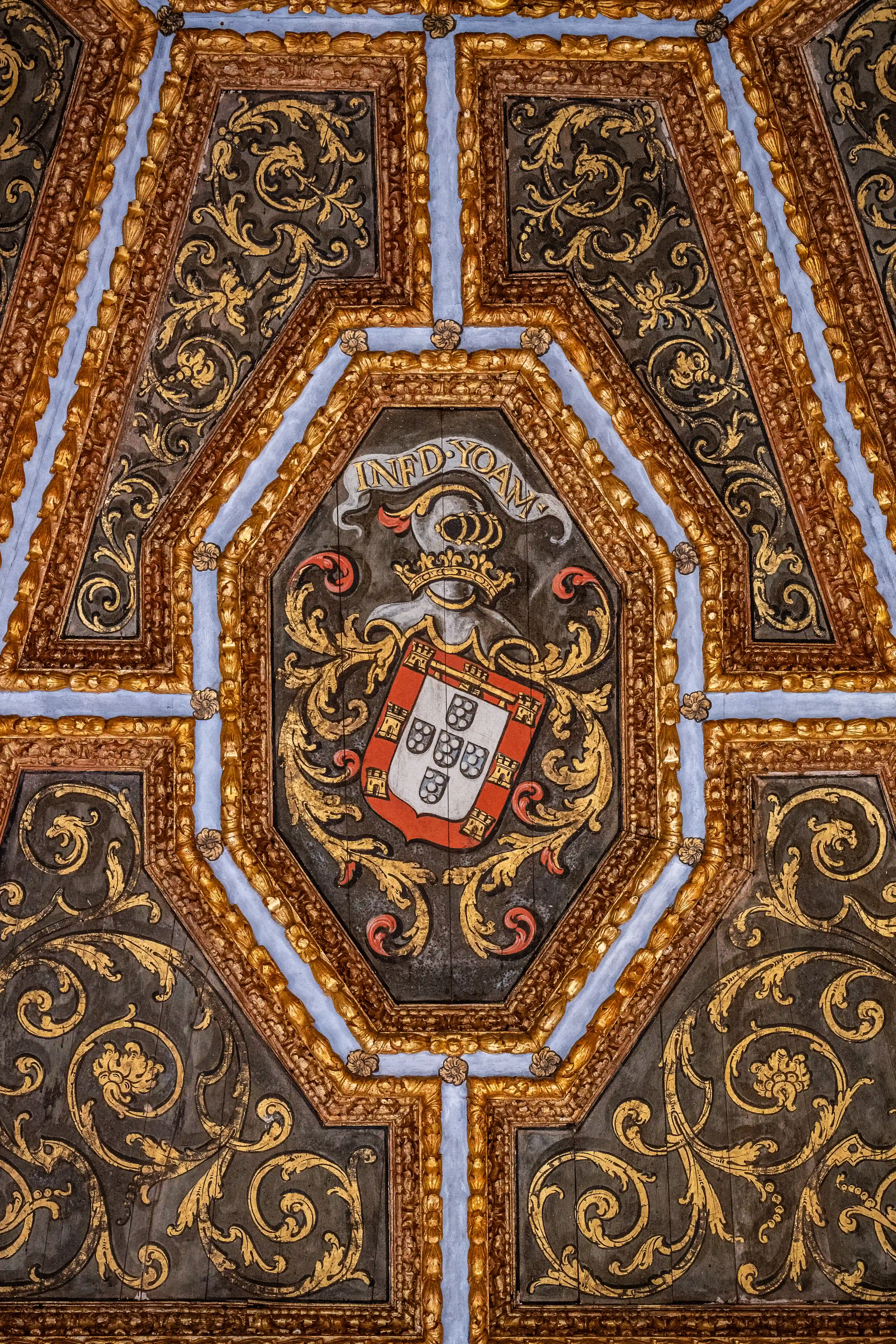 Ornate ceiling panel displaying the Portuguese coat of arms in the centre, surrounded by intricate gold scrollwork on a dark background, framed by carved wooden mouldings and blue detailing.