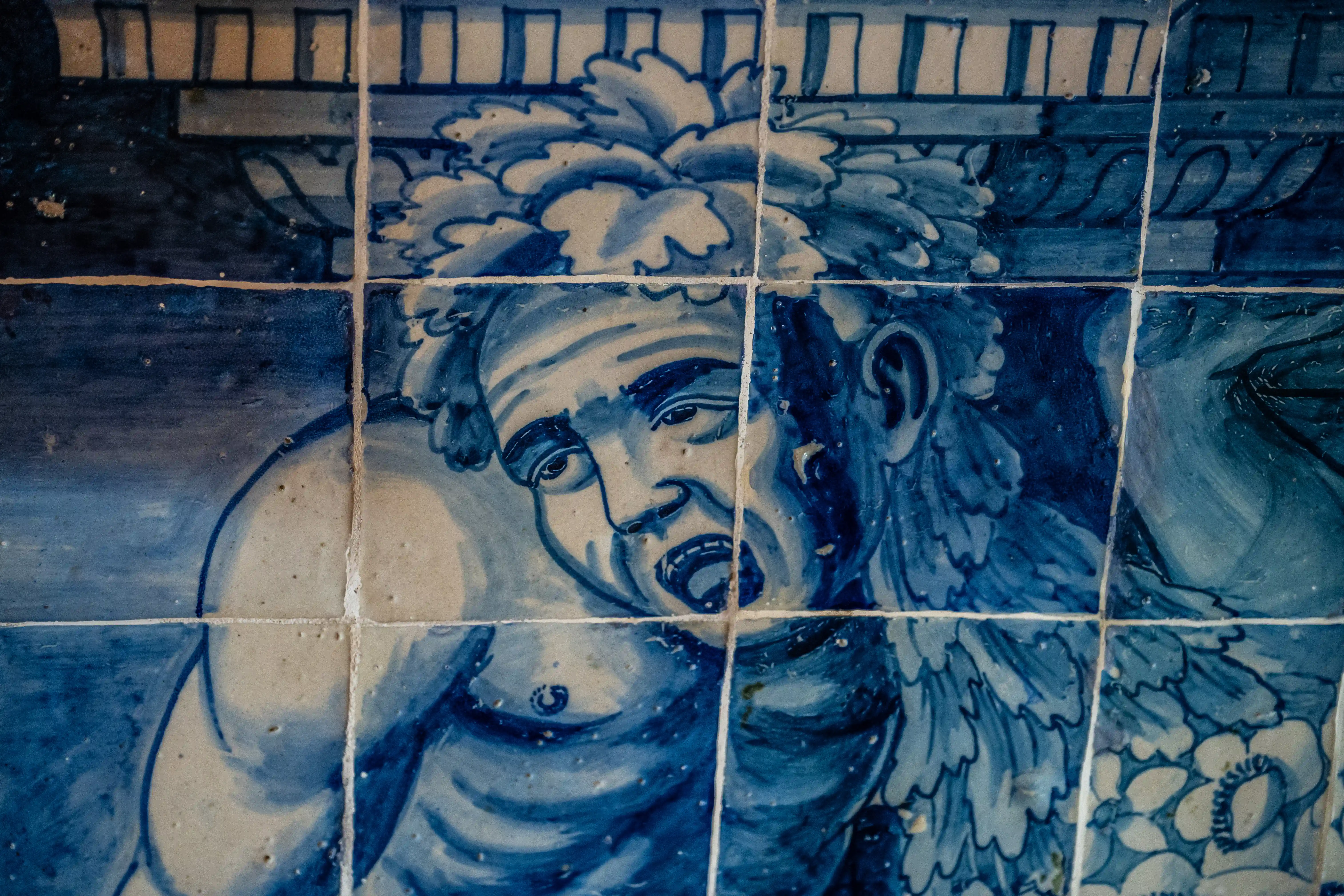 A close-up of a blue and white azulejo tile mural depicting the face and upper body of a mythological figure with a leafy headdress and an open mouth.