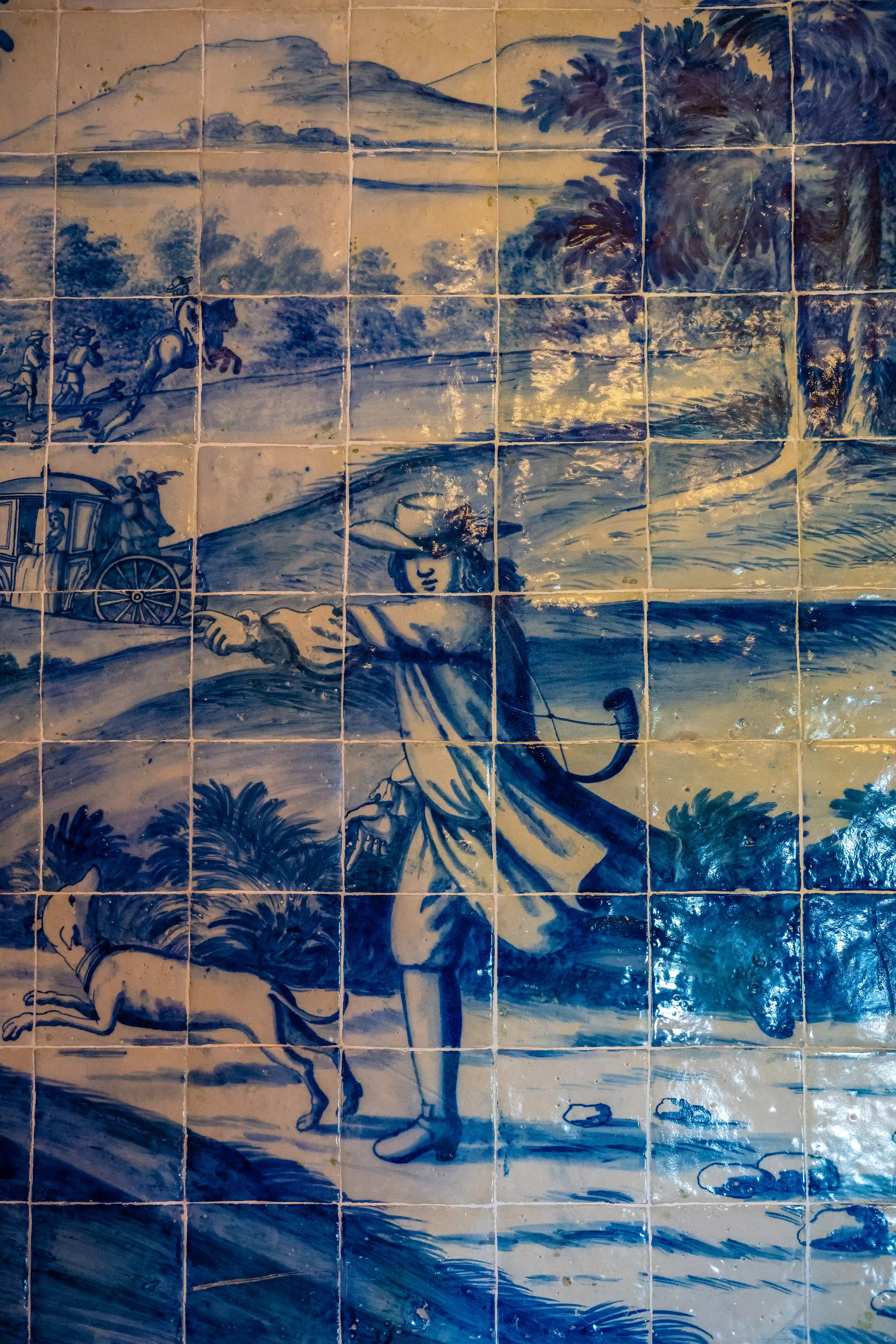  A tiled mural in blue and white depicting a historical scene with a person, a dog, a horse-drawn carriage, and figures on horseback in a landscape.