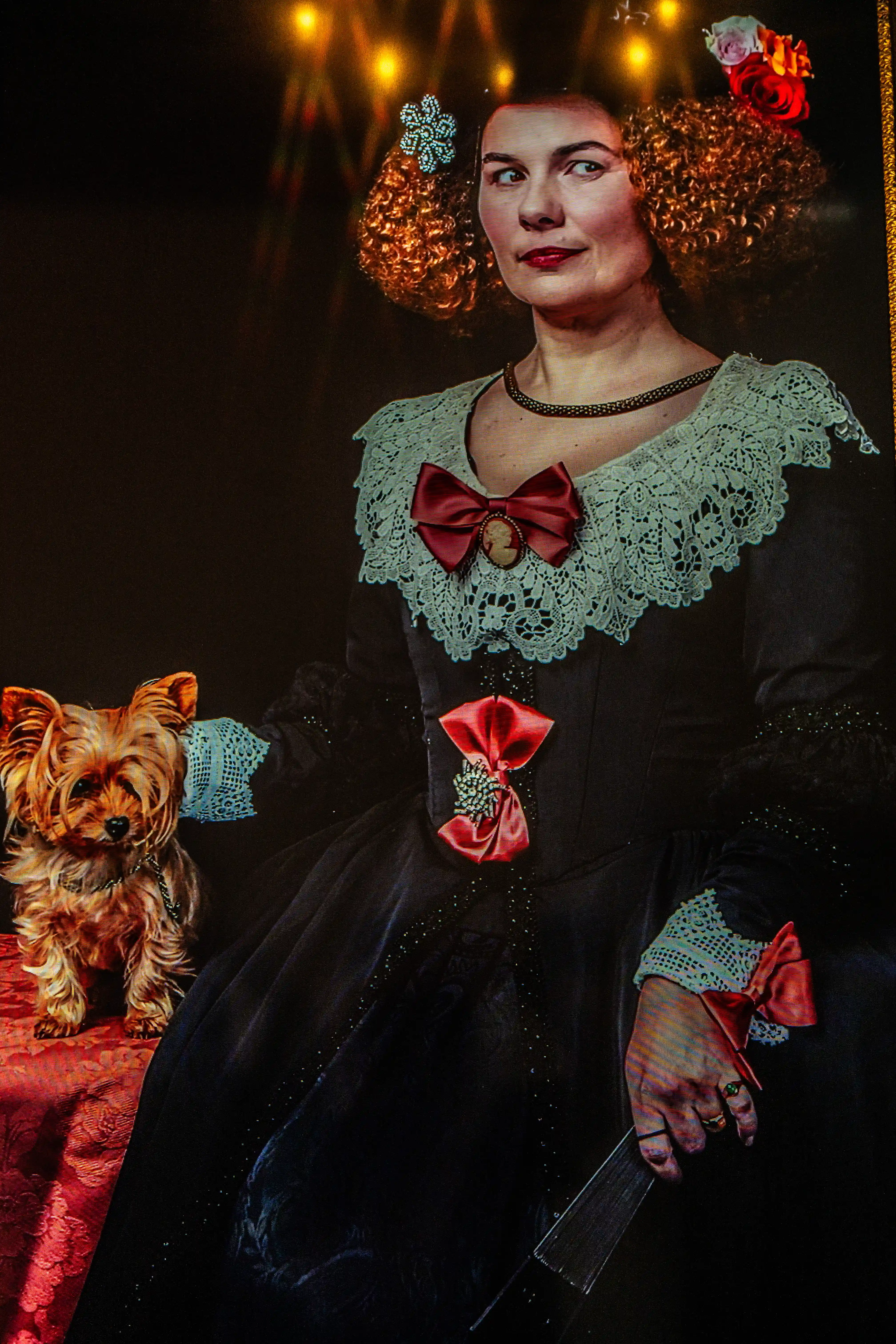 A woman in a black Victorian-style dress with lace and pink bows stands next to a Yorkshire Terrier.