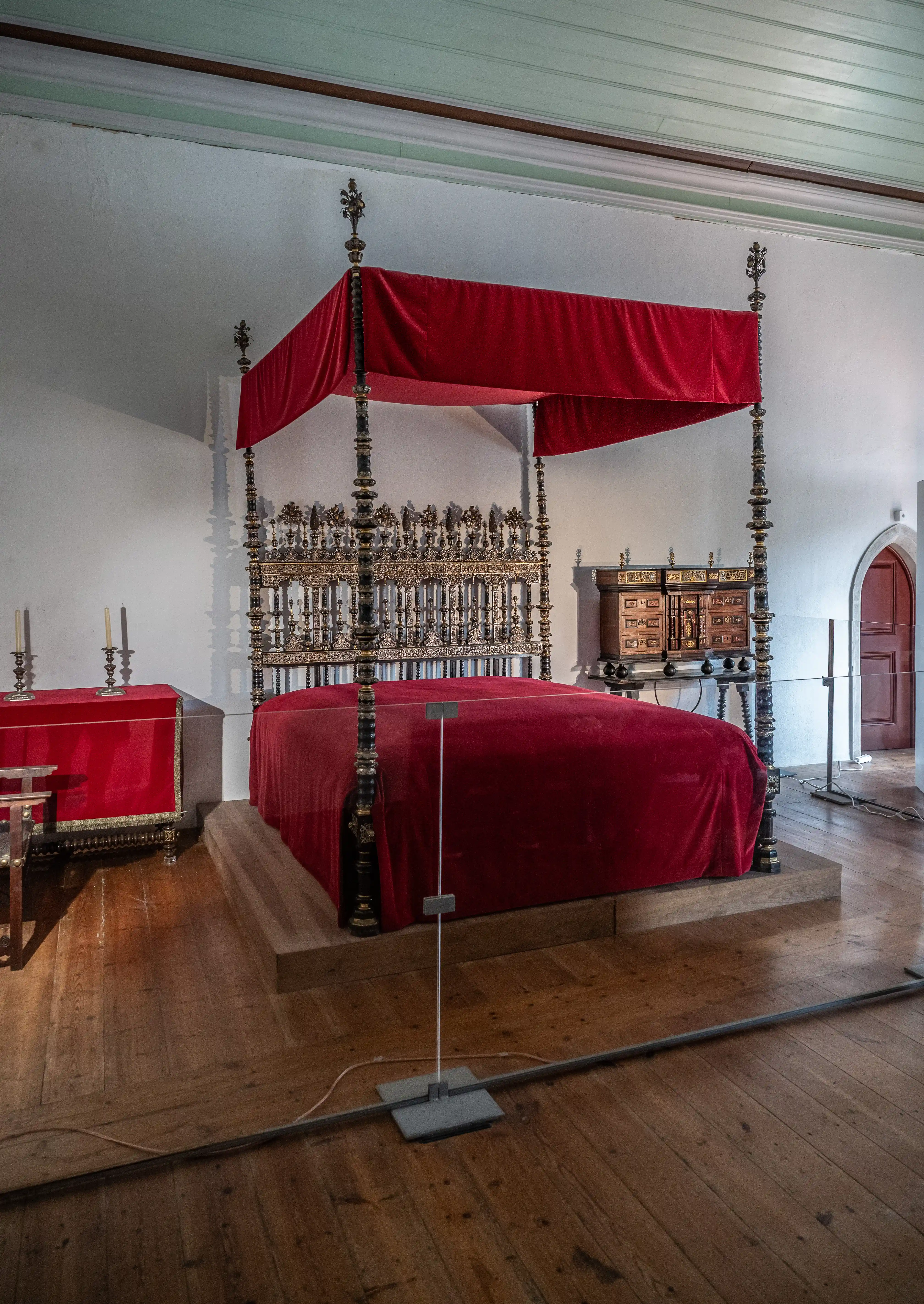 A large, ornate four-poster bed with a red velvet canopy and headboard, set in a room with wooden floors and historical furniture.