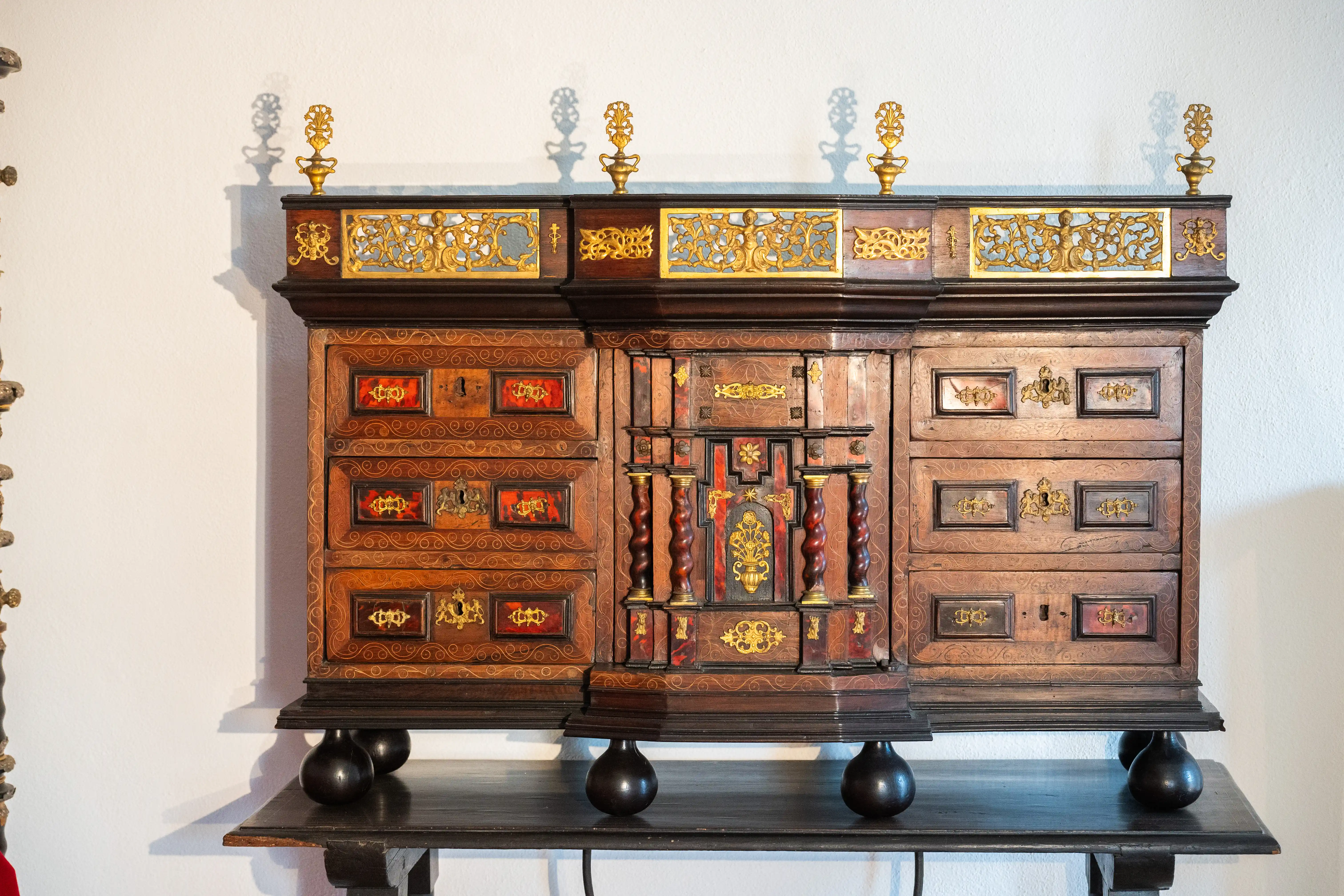 An antique wooden cabinet with multiple drawers and ornate gold detailing, standing on a dark wooden base supported by spherical feet.