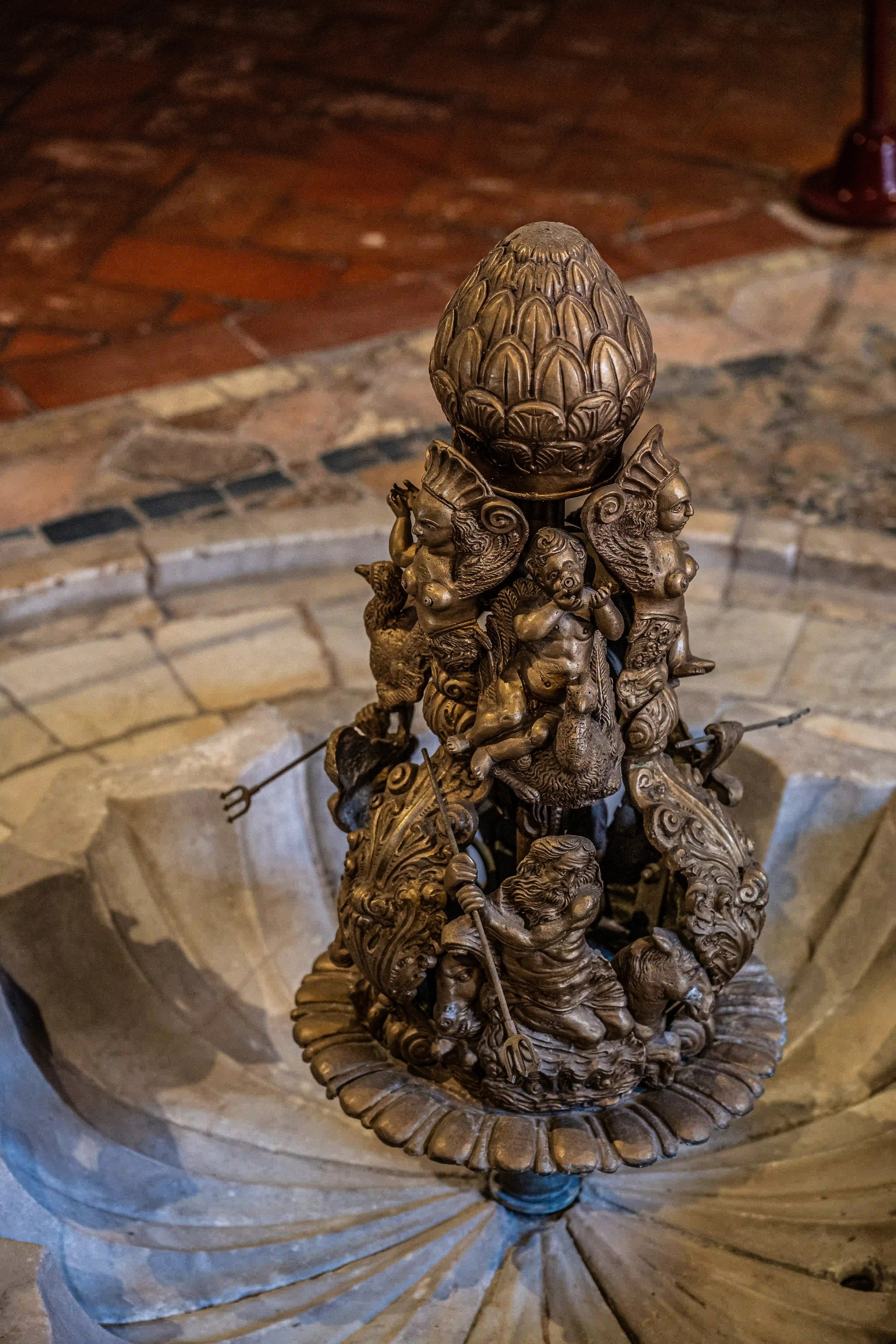 Detailed bronze fountain sculpture with multiple cherubs holding tridents, topped with an artichoke-like finial, set in a stone basin.