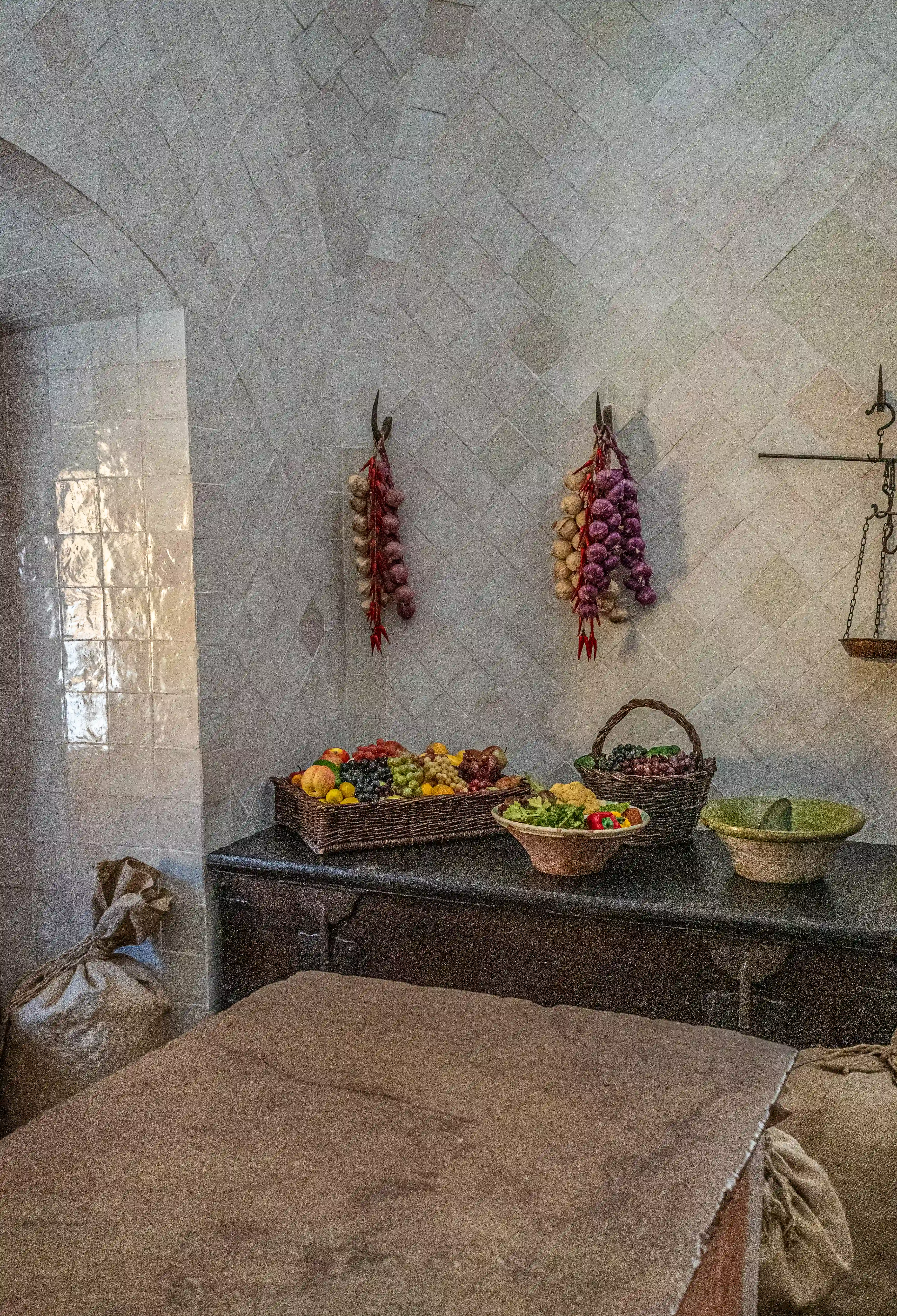 A rustic kitchen interior with arched, tiled walls. Bunches of red onions and garlic hang from hooks. A dark wooden counter displays a basket of mixed fruit, a bowl of salad, and a green bowl. Sacks of grain are piled in the foreground, and a rough stone surface dominates the lower part of the image.