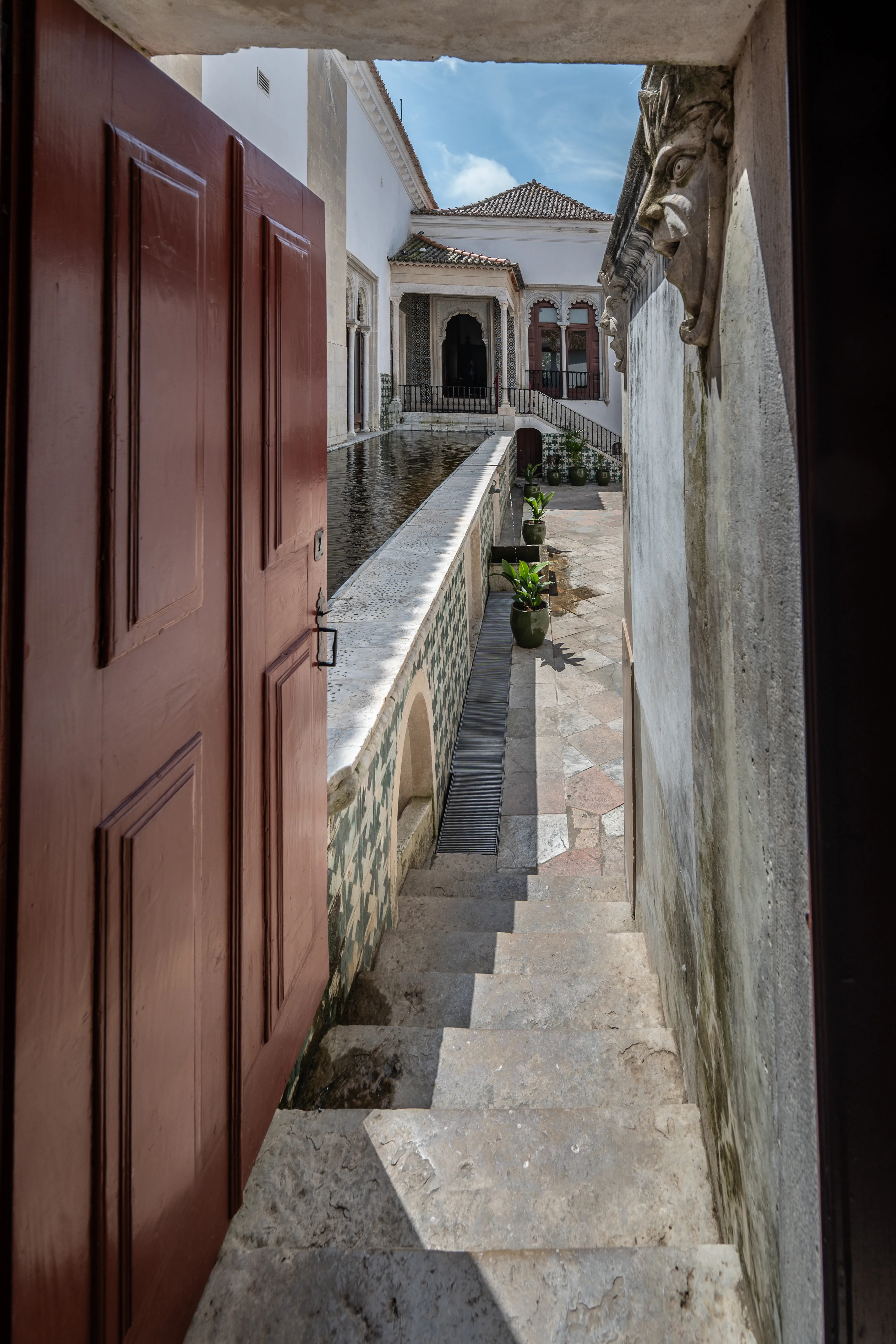 View from inside a dark doorway looking out into a sunny courtyard with a water feature, tiled walls, and stone stairs.