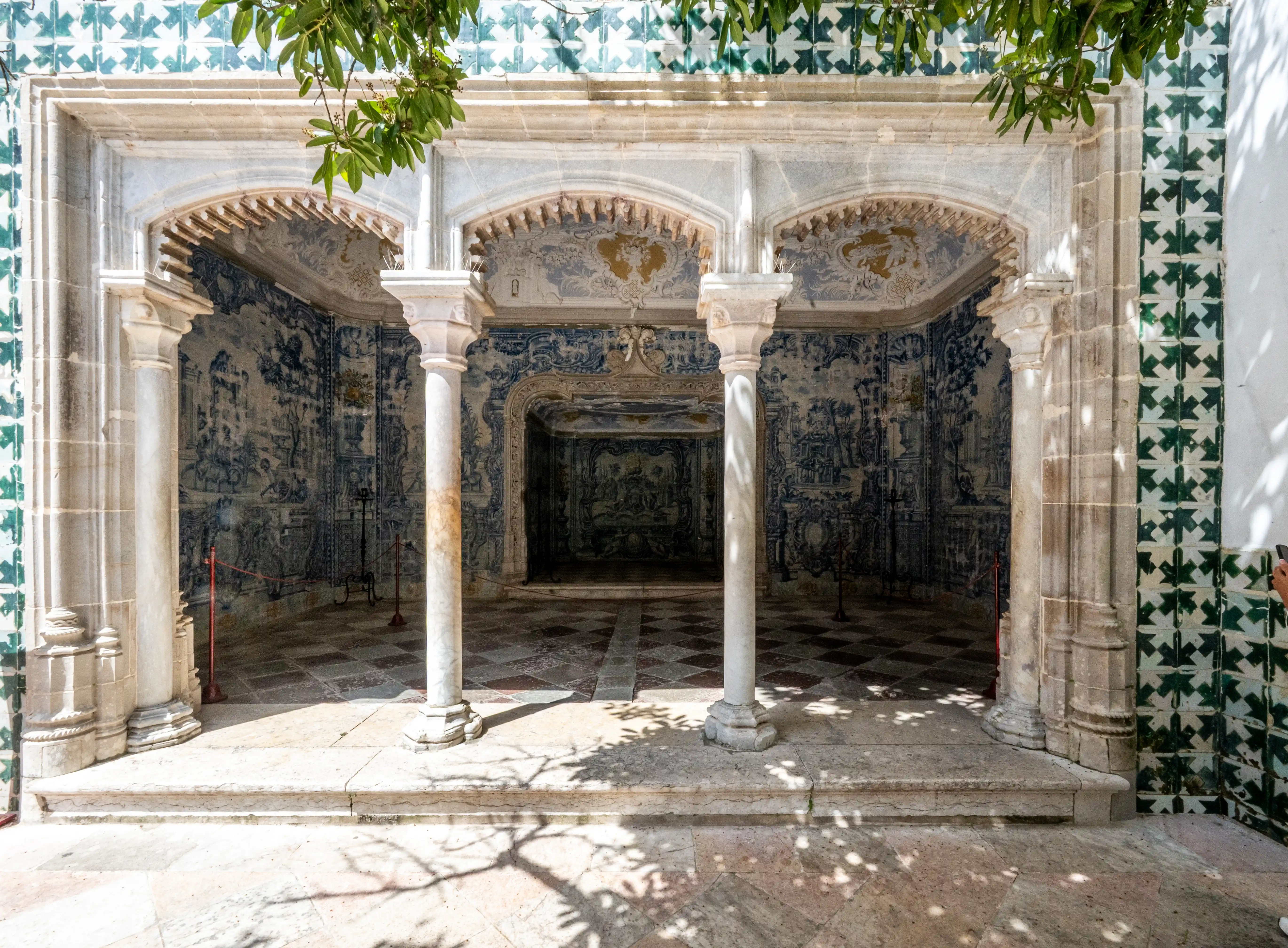 An ornate archway adorned with blue and white azulejo tiles, with sunlight casting shadows on the floor and foliage overhead.