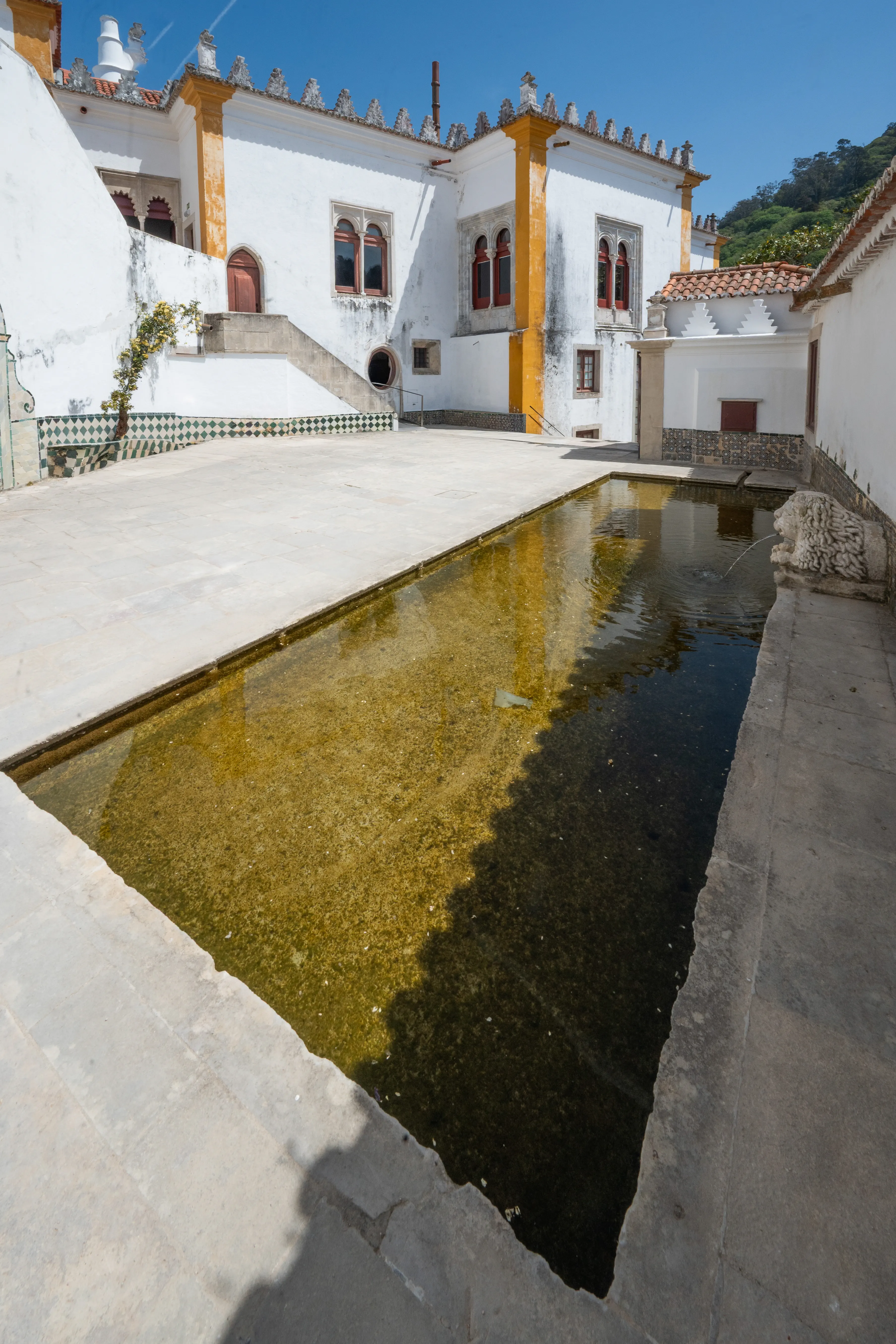 A woman in a light-coloured outfit takes a selfie in the courtyard of a European palace, with a rectangular pool of water reflecting the blue sky and surrounding white buildings.