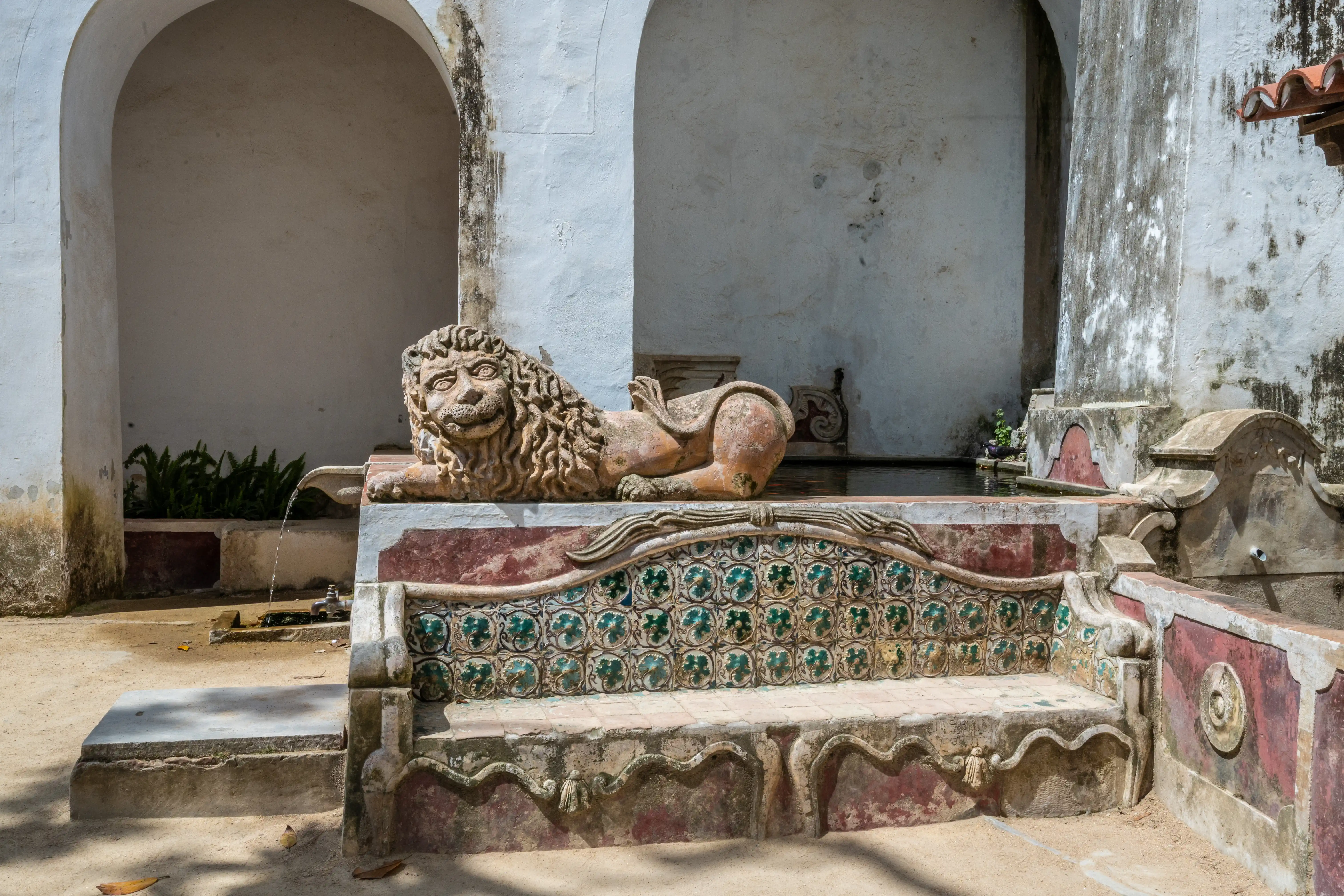 A stone lion sculpture with a flowing mane rests on the edge of a fountain, overlooking a tiled bench. White arched walls and a sandy courtyard complete the scene.