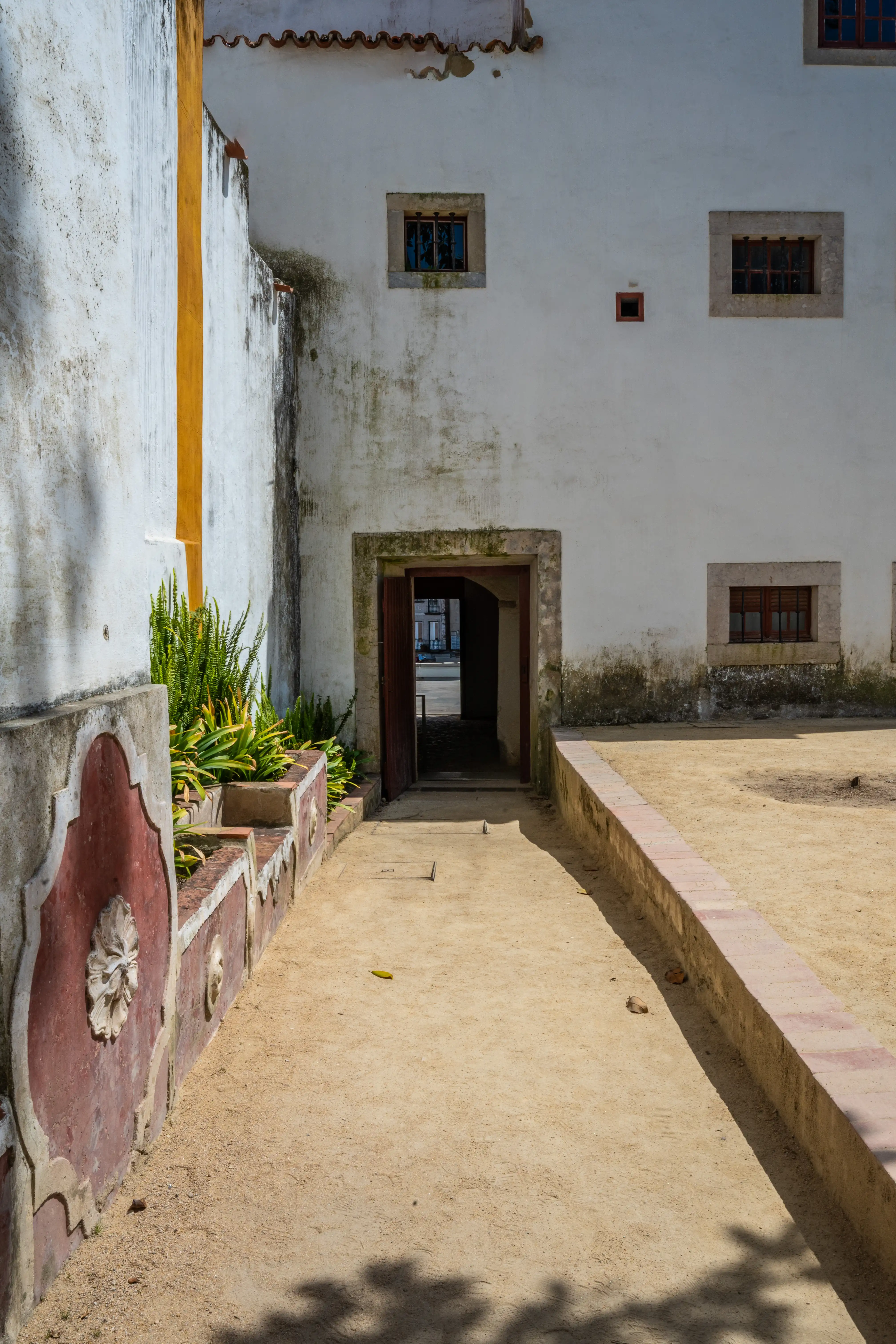 A view down a sandy courtyard towards an open doorway of a white building. A decorative planter with greenery is on the left, and shadows from trees are cast on the ground.