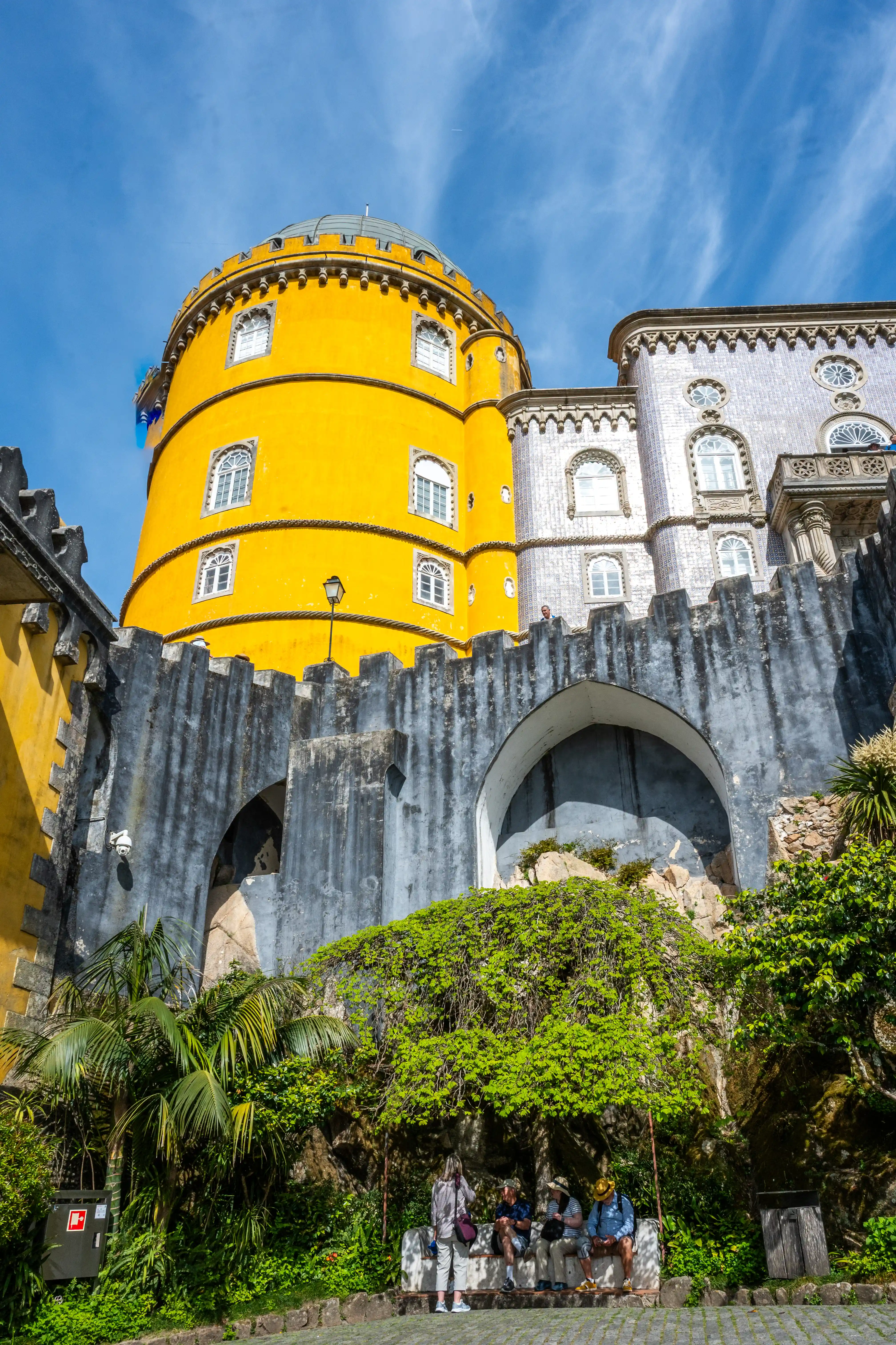 A bright yellow tower and a building adorned with blue and white tiles, part of Pena Palace, stand against a partly cloudy blue sky. In the foreground, a stone wall is covered with dense green foliage, with several people sitting on a bench beneath the shade of trees.