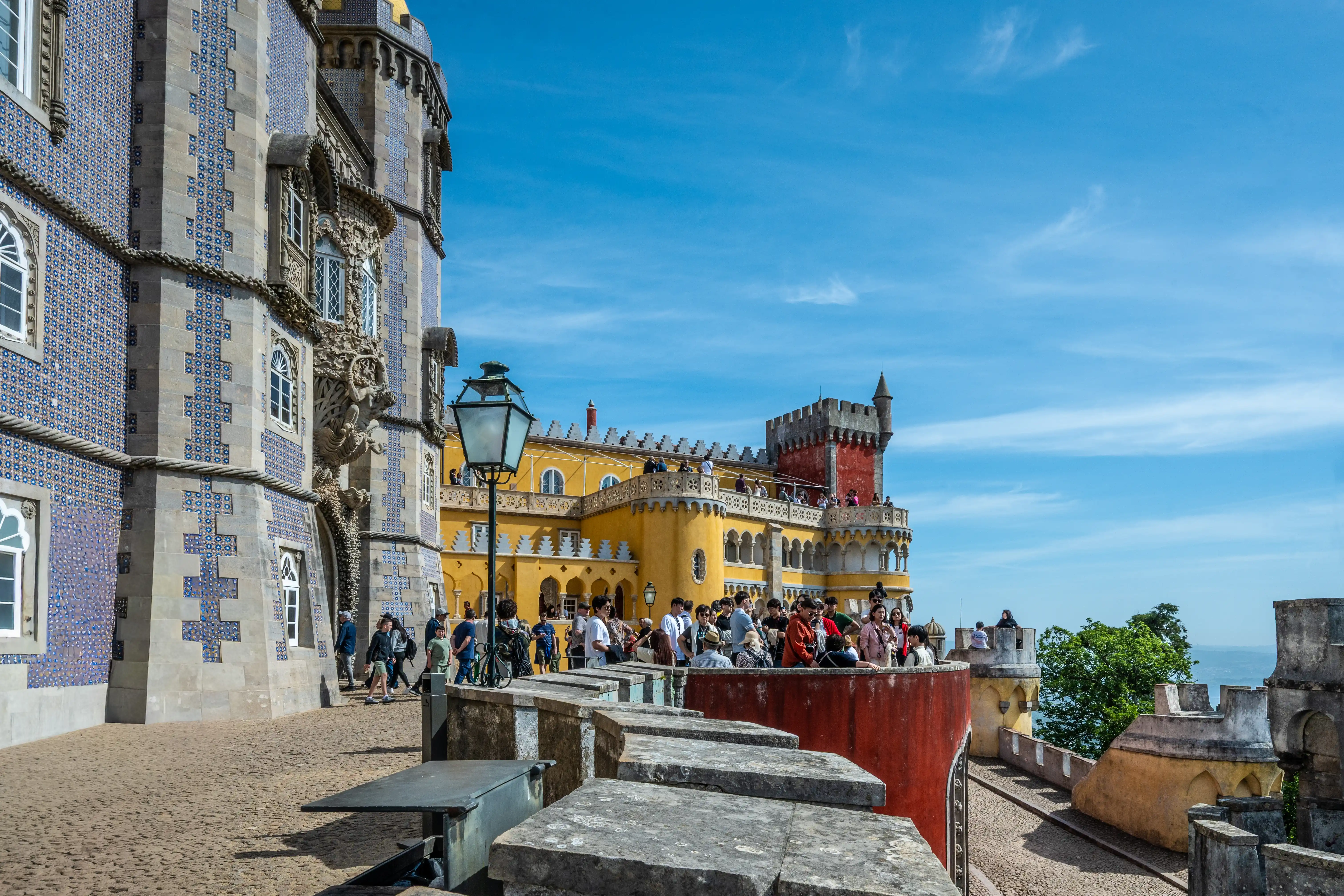 he ornate, colourful Pena Palace in Sintra, Portugal, under a bright blue sky with scattered clouds. The palace has a section covered in blue and white patterned tiles and another section painted bright yellow with a red tower. Many tourists are visible on the balconies and pathways.