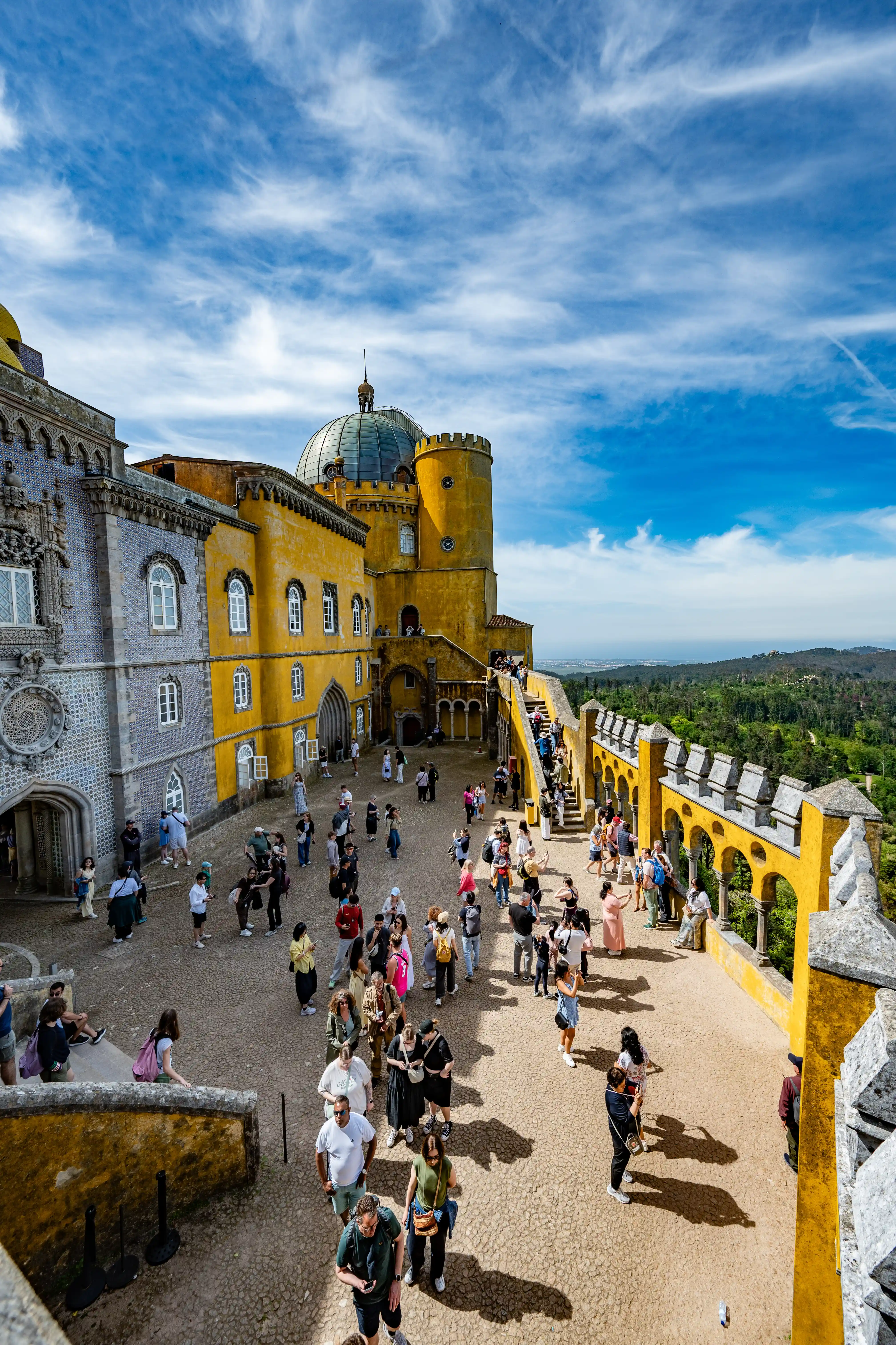 A crowd of tourists mills about the courtyard of a yellow and grey palace with a blue dome, set against a backdrop of green hills and a partly cloudy sky.
