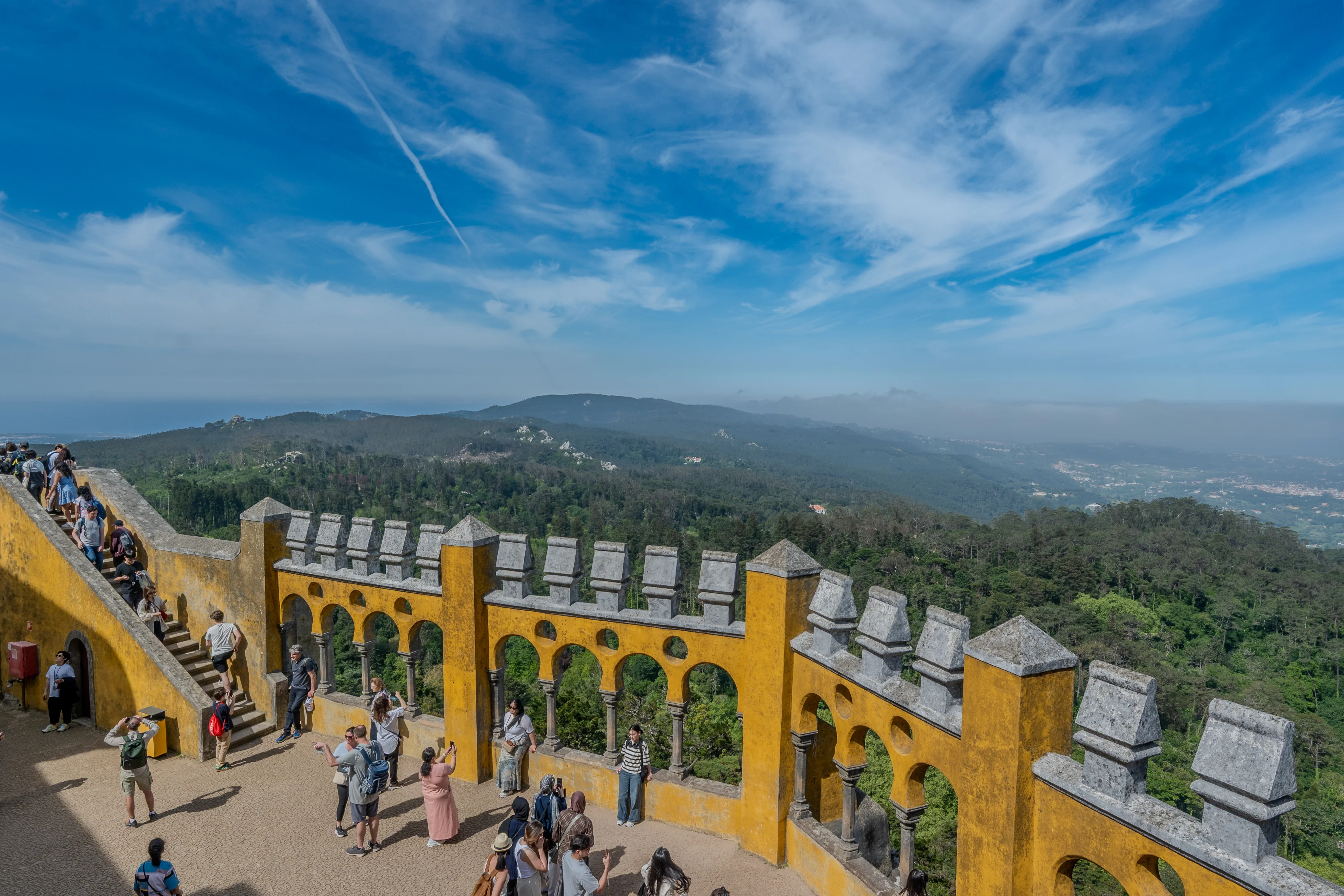 A crowd of tourists walks up the yellow stone steps of a castle courtyard, with a vast green landscape and distant city stretching out below a cloudy blue sky.
