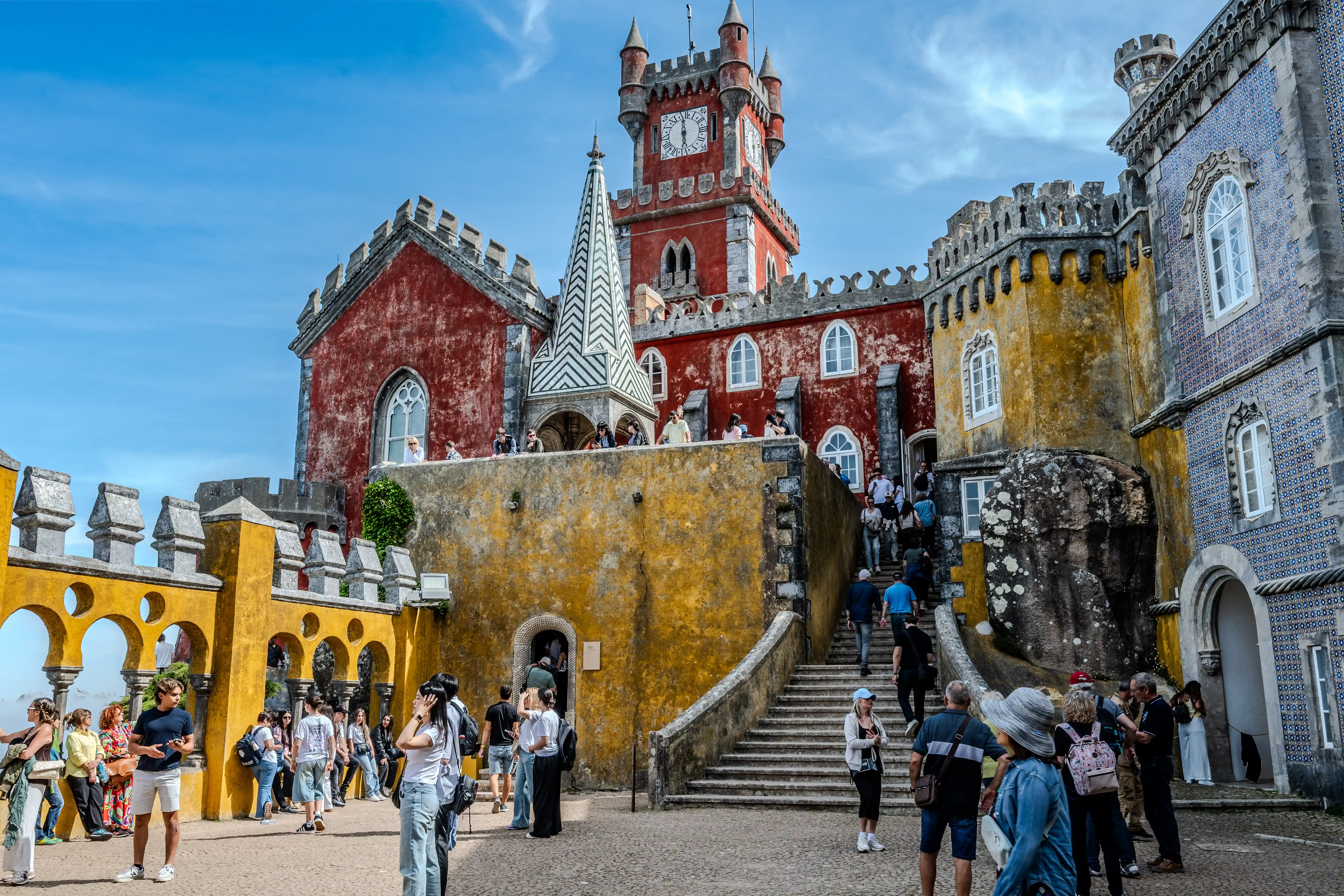 A wide shot of the colourful Pena Palace, with many tourists walking around the courtyards and up the grand stone staircase. The bright red and yellow buildings are adorned with decorative turrets, arched windows, and patterned tiling.