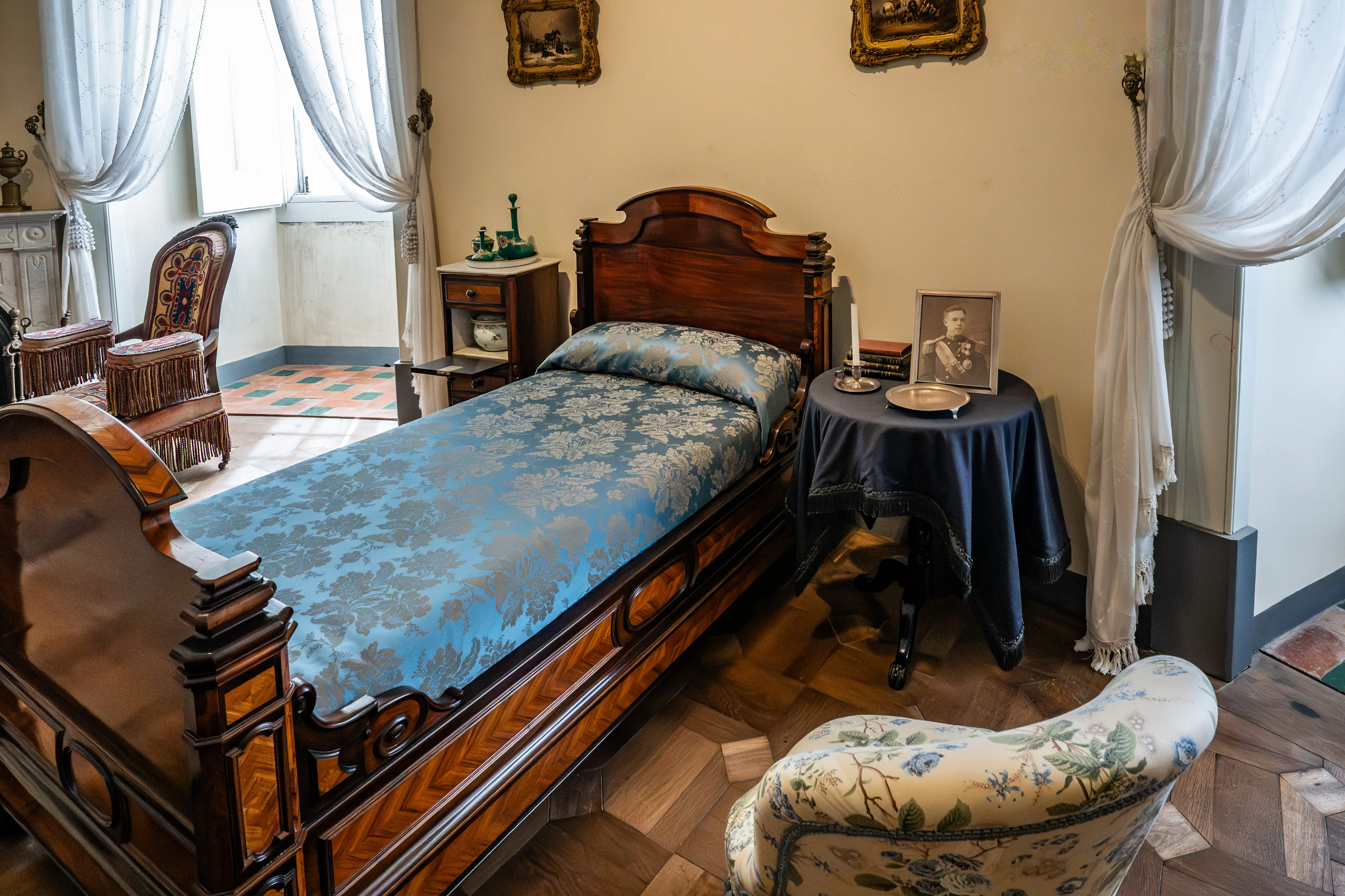 A vintage bedroom scene with a large, ornate wooden bed covered in a blue patterned duvet. To the right of the bed, a small round table has a framed portrait of a man in uniform, a stack of books, and a silver candlestick. A dark wood bedside table with decorative bottles sits to the left of the bed. Two antique chairs with fringed upholstery are visible near a fireplace and a window with sheer white curtains.
