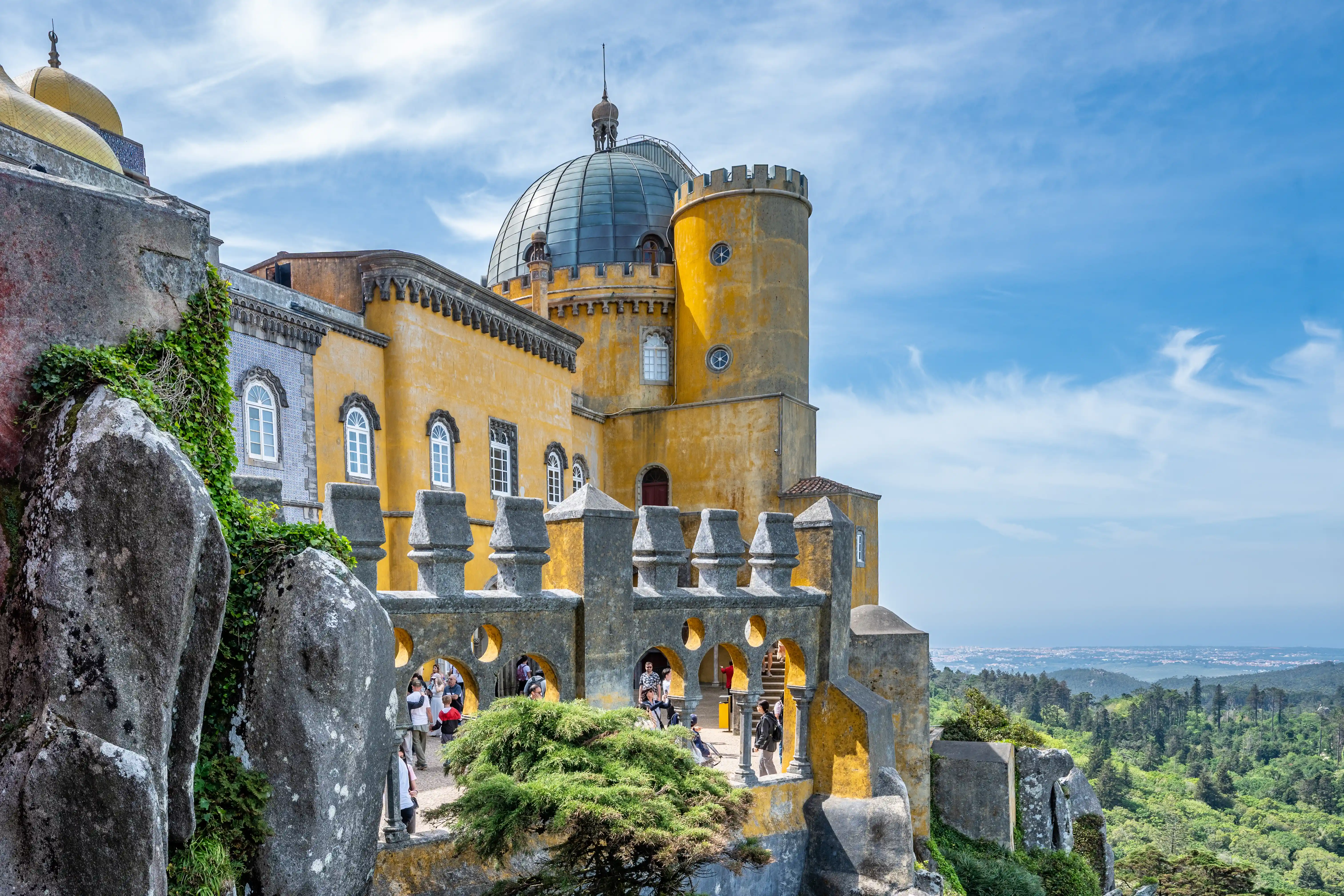 A brightly coloured yellow palace with a large dome and a cylindrical tower, perched on a rocky cliff surrounded by dense green woodland. People are visible on the palace terraces.