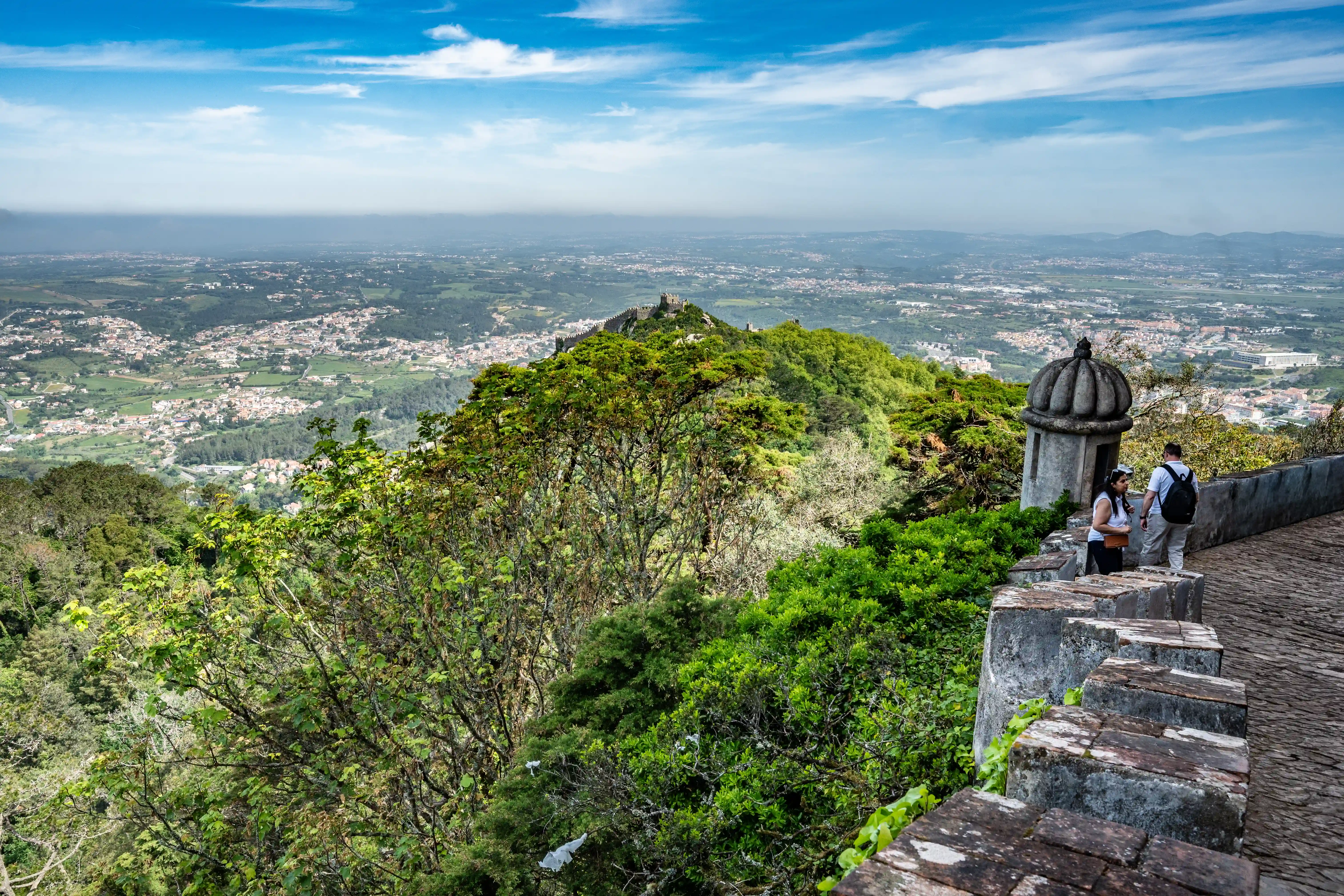 Two tourists stand on a stone pathway, looking out at a vast landscape dominated by the Castle of the Moors on a green hilltop, with a town spread out below under a cloudy blue sky.