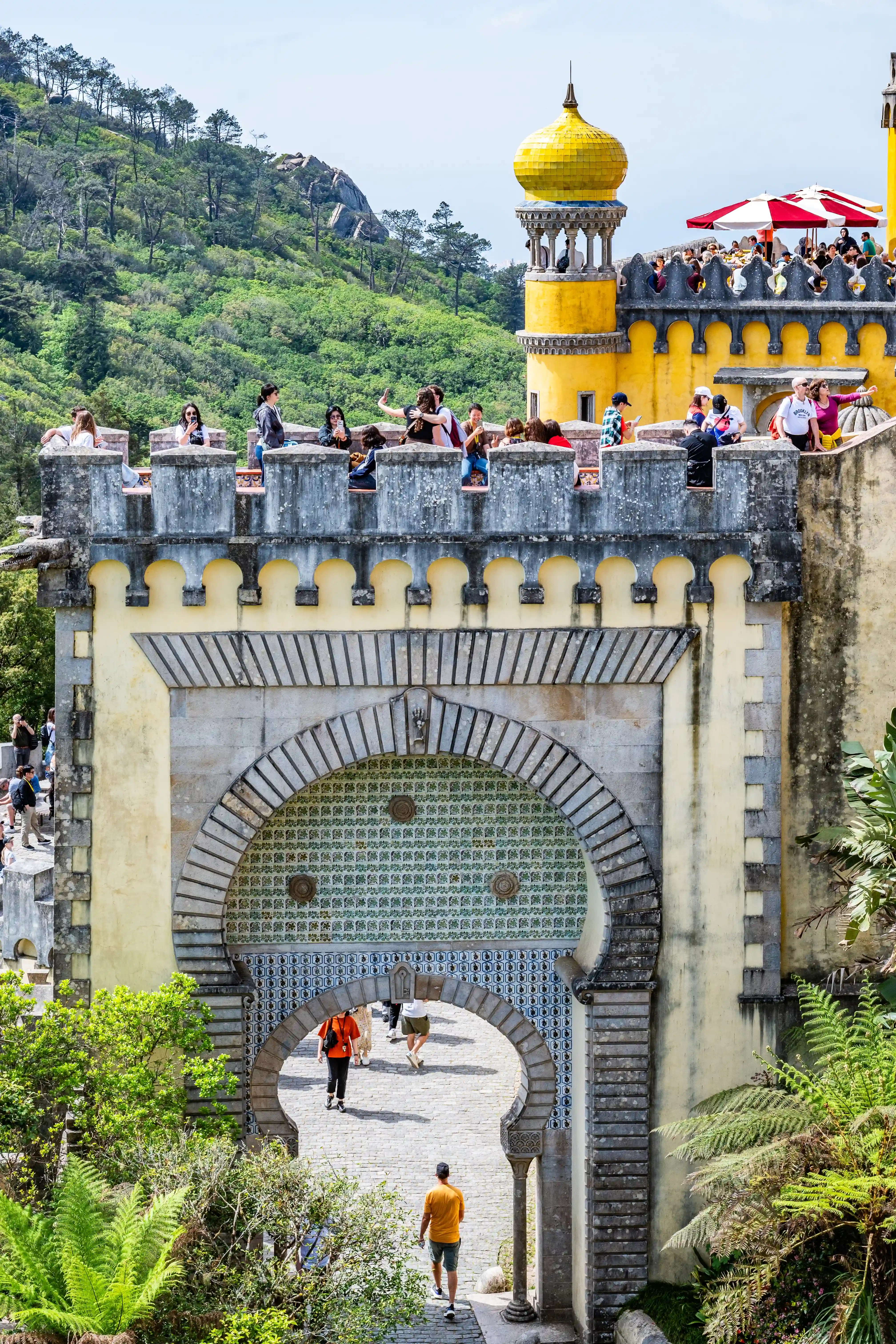 A group of tourists on the battlements of a yellow palace with a golden dome, overlooking a forested hillside