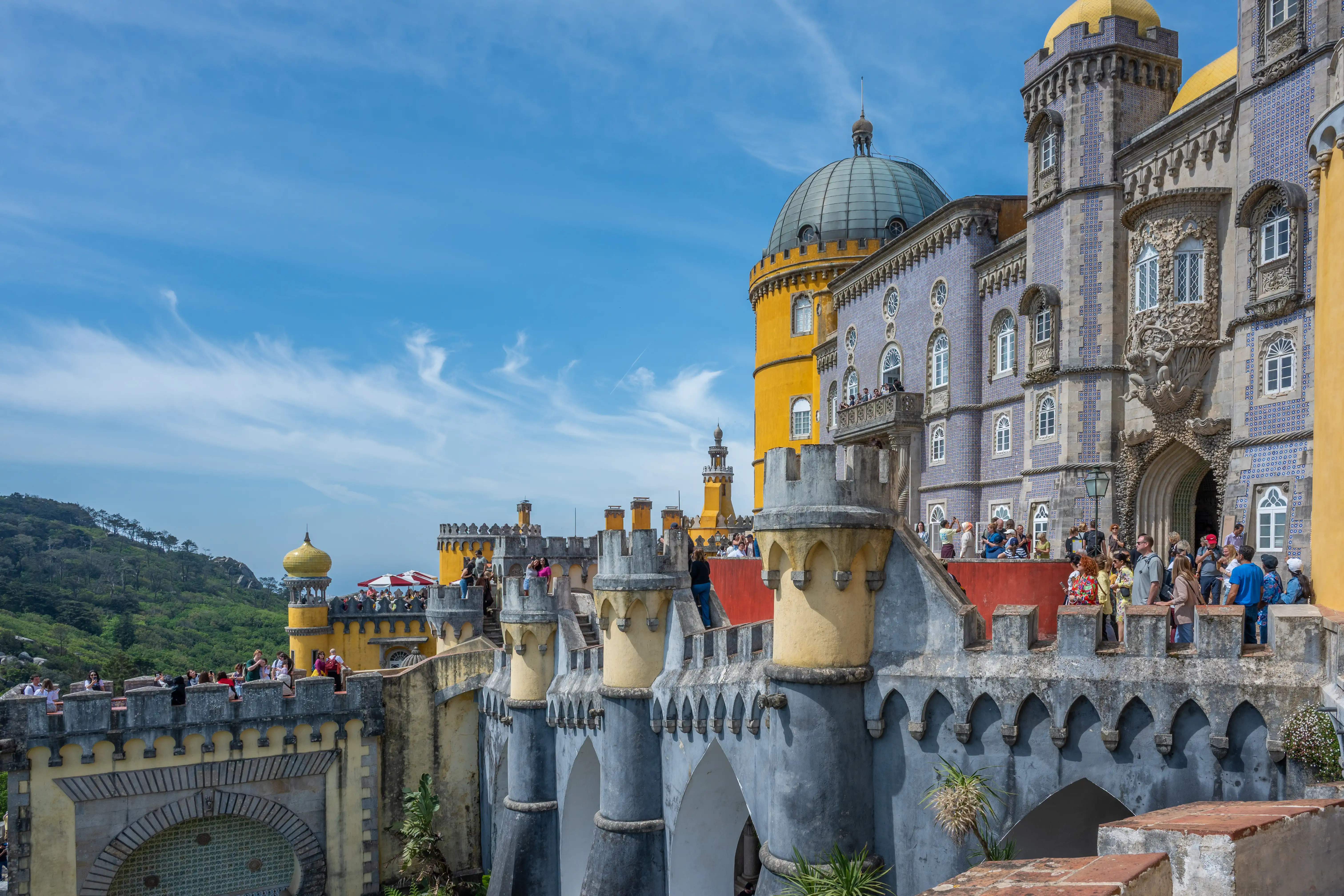 The exterior of the vibrant Pena Palace in Sintra, Portugal, featuring yellow and blue facades, ornate architectural details, and a domed tower, with several tourists visible on its terraces.