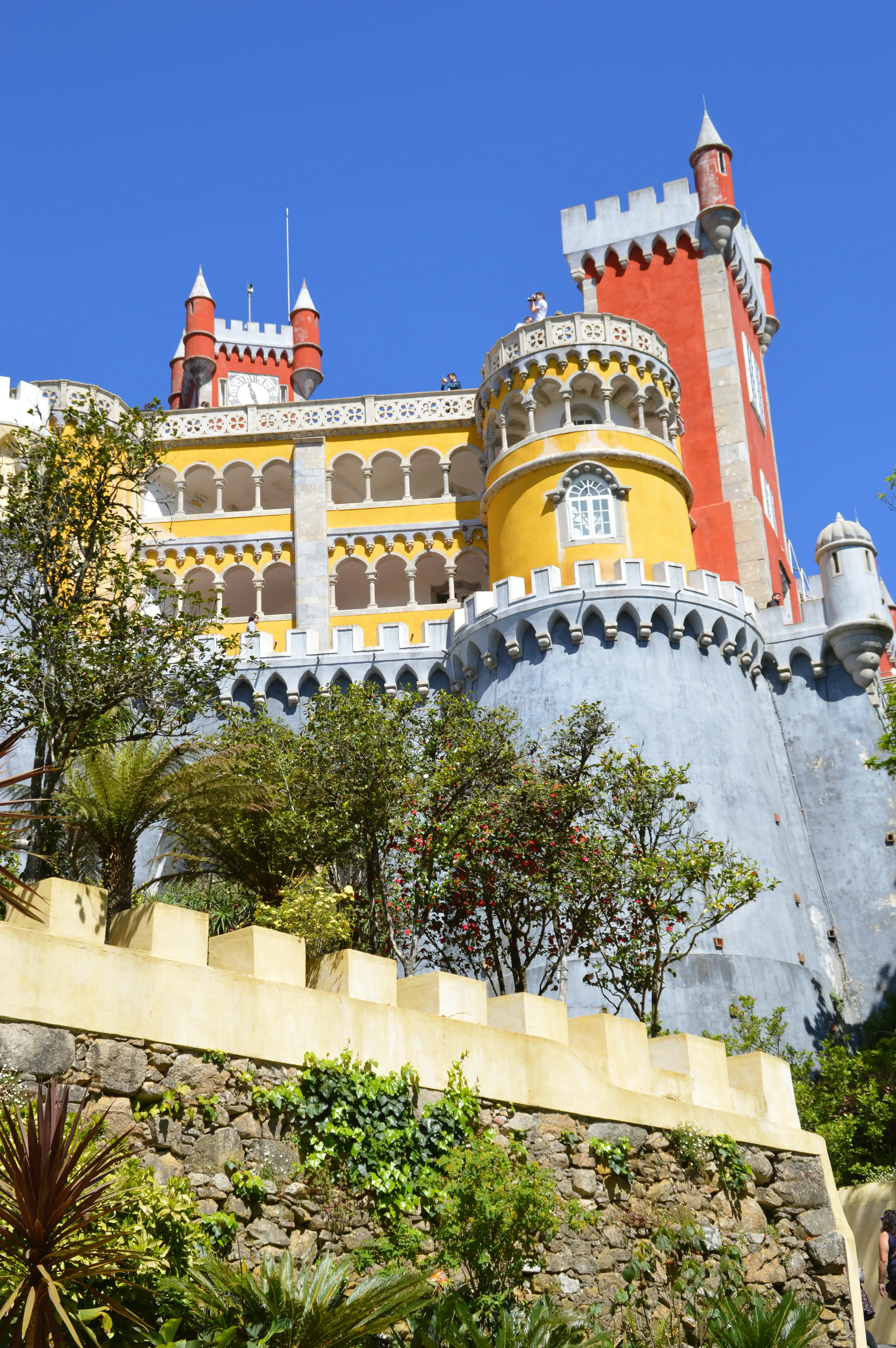 A low-angle shot of the Pena Palace, a historic monument with red and yellow towers, under a clear blue sky. Green foliage and a stone wall with crenellations are visible in the foreground.