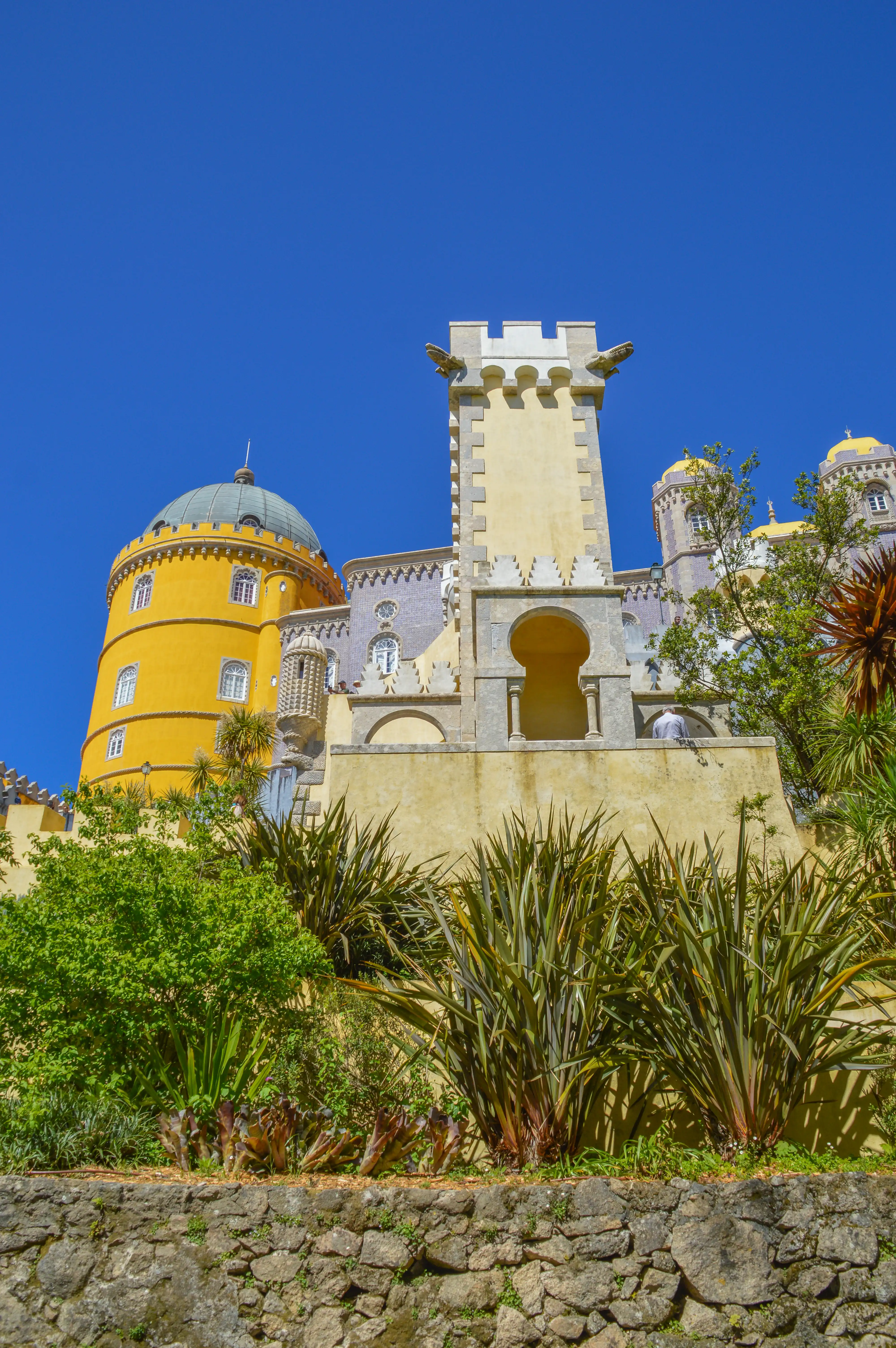A vibrant yellow tower with a green dome, part of Pena Palace, is seen against a bright blue sky, with decorative gargoyles on a crenellated turret and lush green vegetation in the foreground.