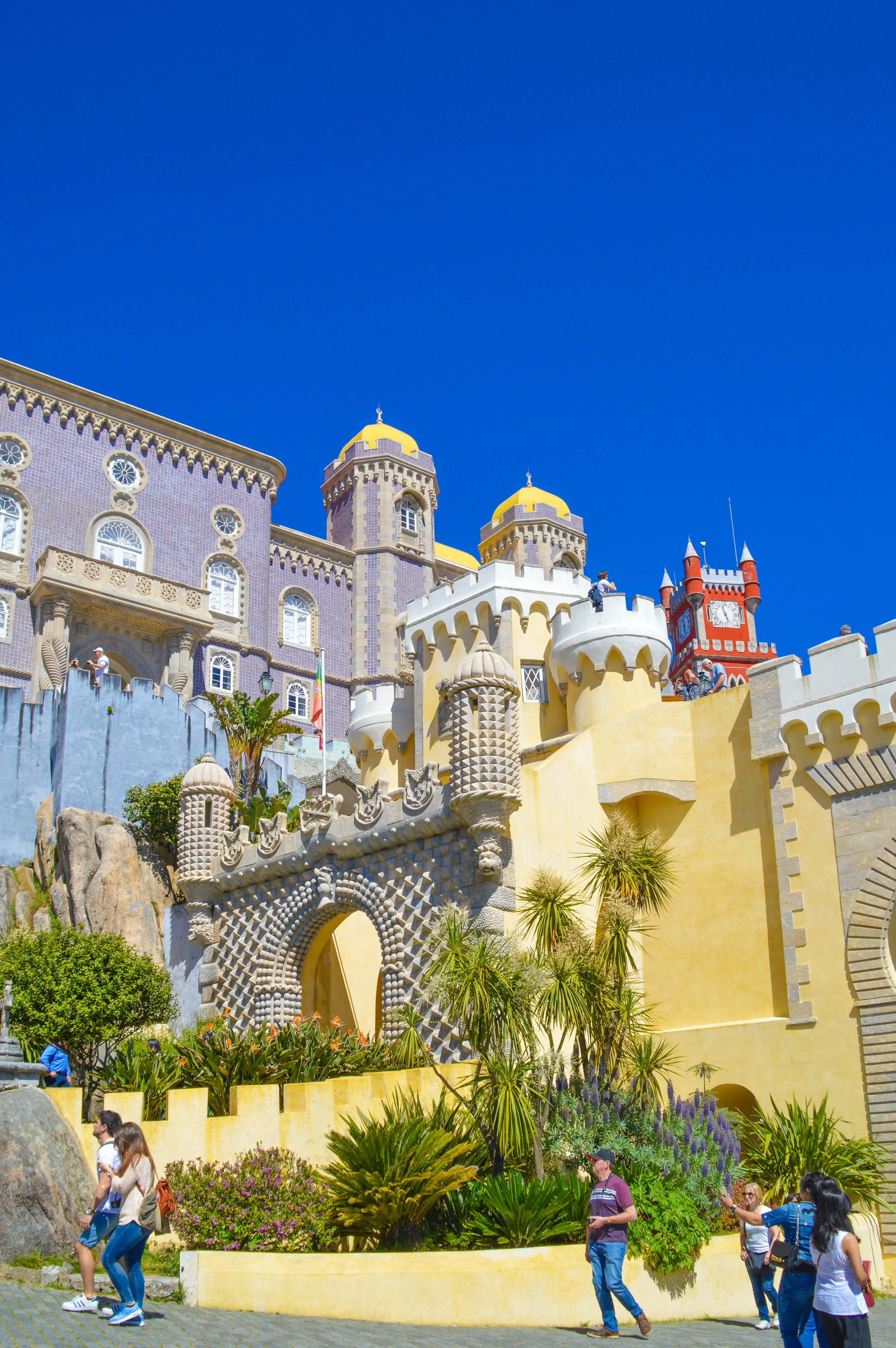 A wide-angle shot of the Pena Palace in Sintra, Portugal, under a bright blue sky. The palace features striking yellow and blue facades, with ornate towers, domes, and decorative stonework. Several palm trees and other greenery are visible in the foreground, and a few tourists are walking on the paved pathway in front of the palace walls.