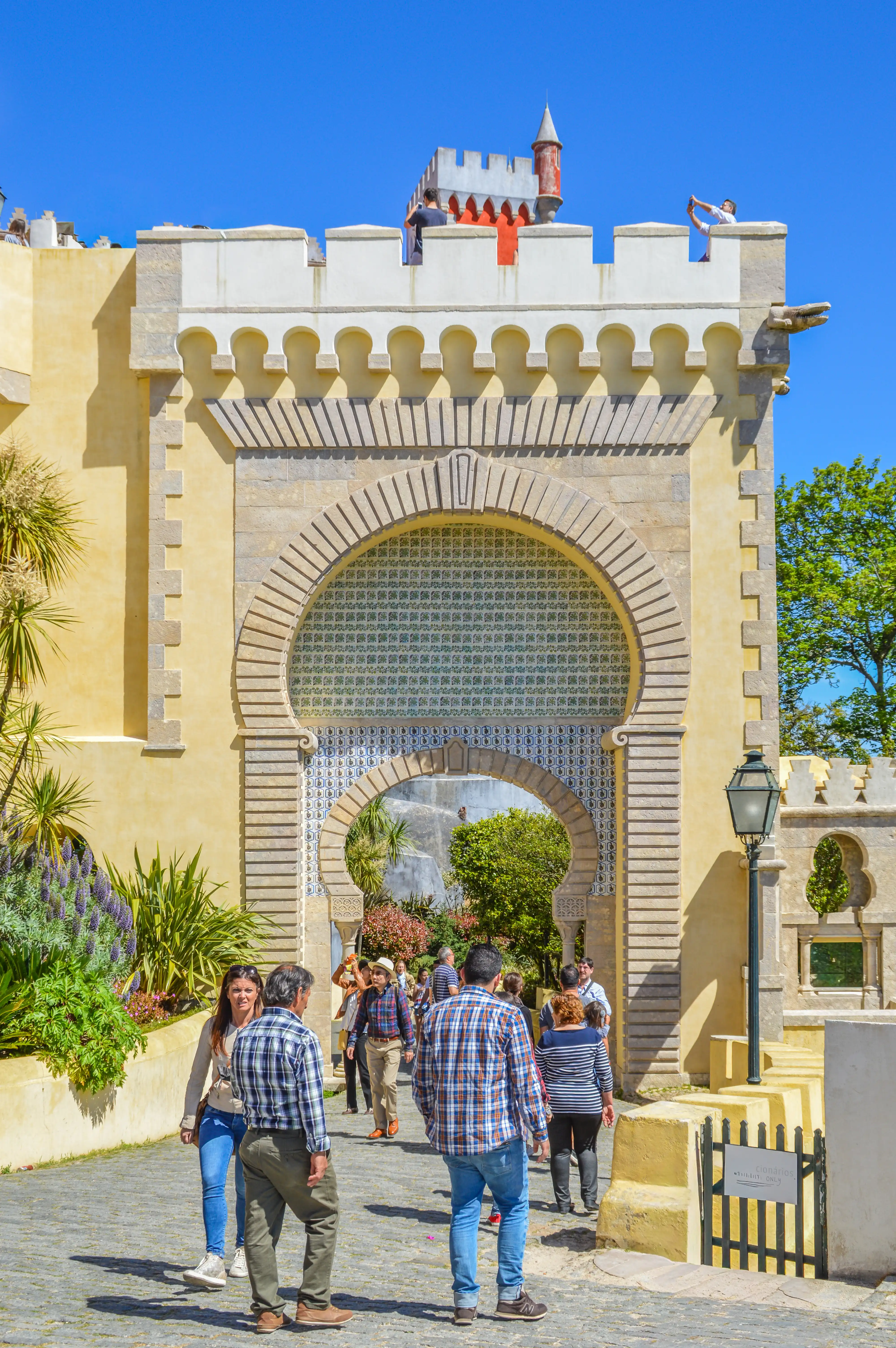  A group of people stroll through a large, colourful, arched entranceway of a palace, with people visible on the ramparts above under a clear blue sky.