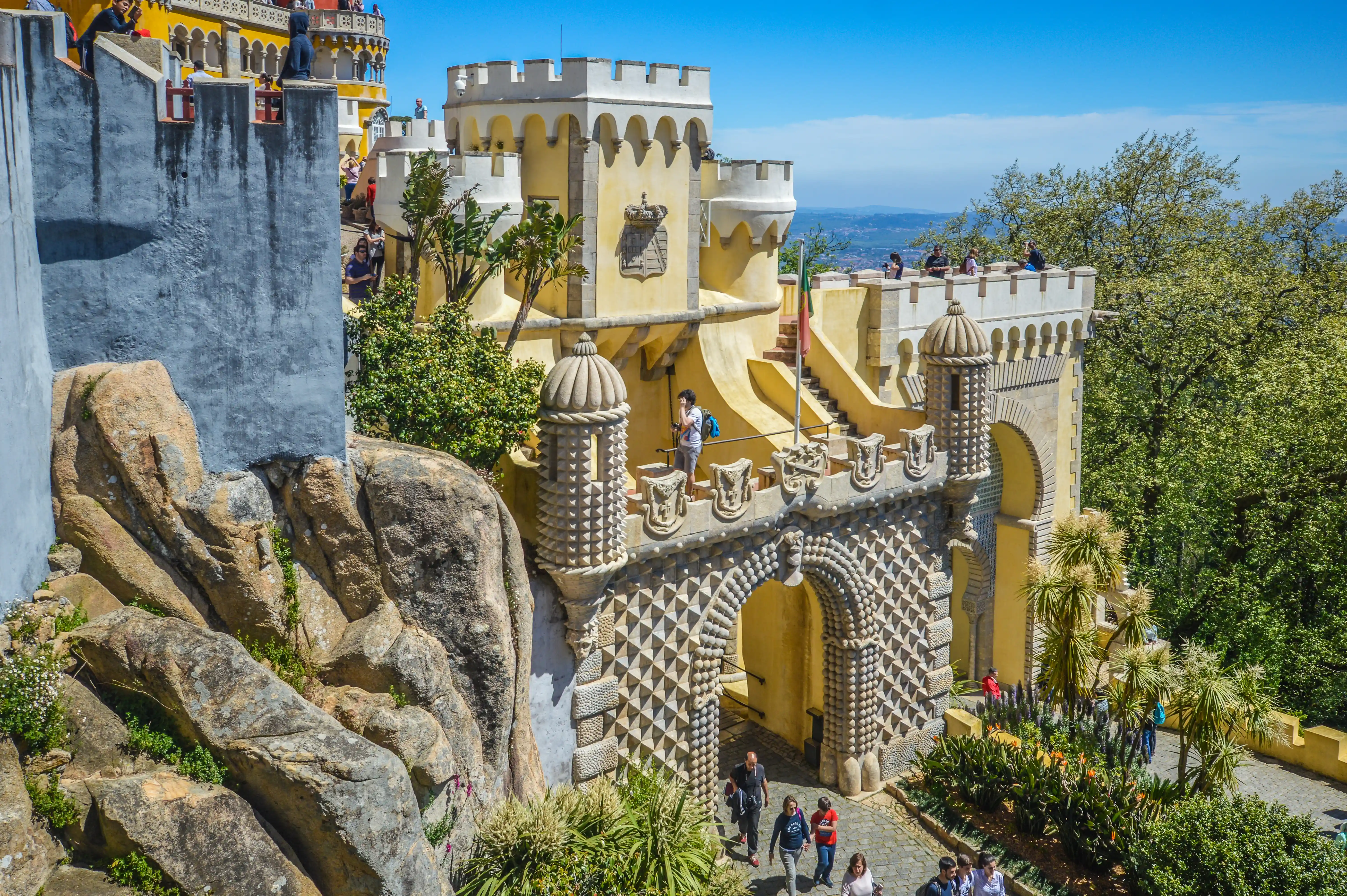 Pena Palace in Sintra, Portugal, with its distinctive yellow and white turrets, arched gateways, and elaborate stonework, is set against a bright blue sky. Green trees and rocks frame the foreground, and several tourists are visible on the pathways and balconies.