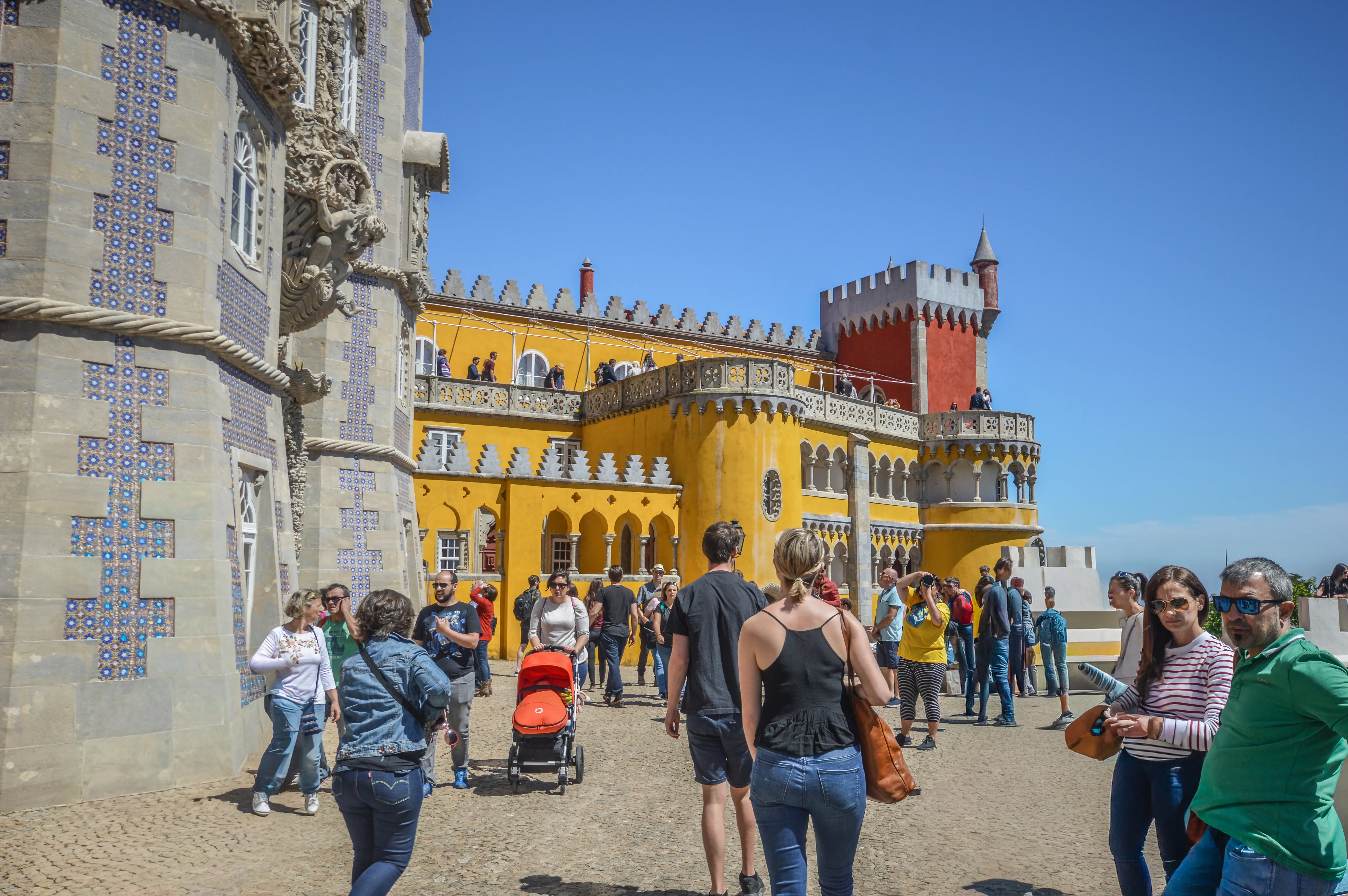 Tourists gathered in the courtyard of the Pena Palace on a sunny day, with the yellow and red palace buildings and blue sky in the background.