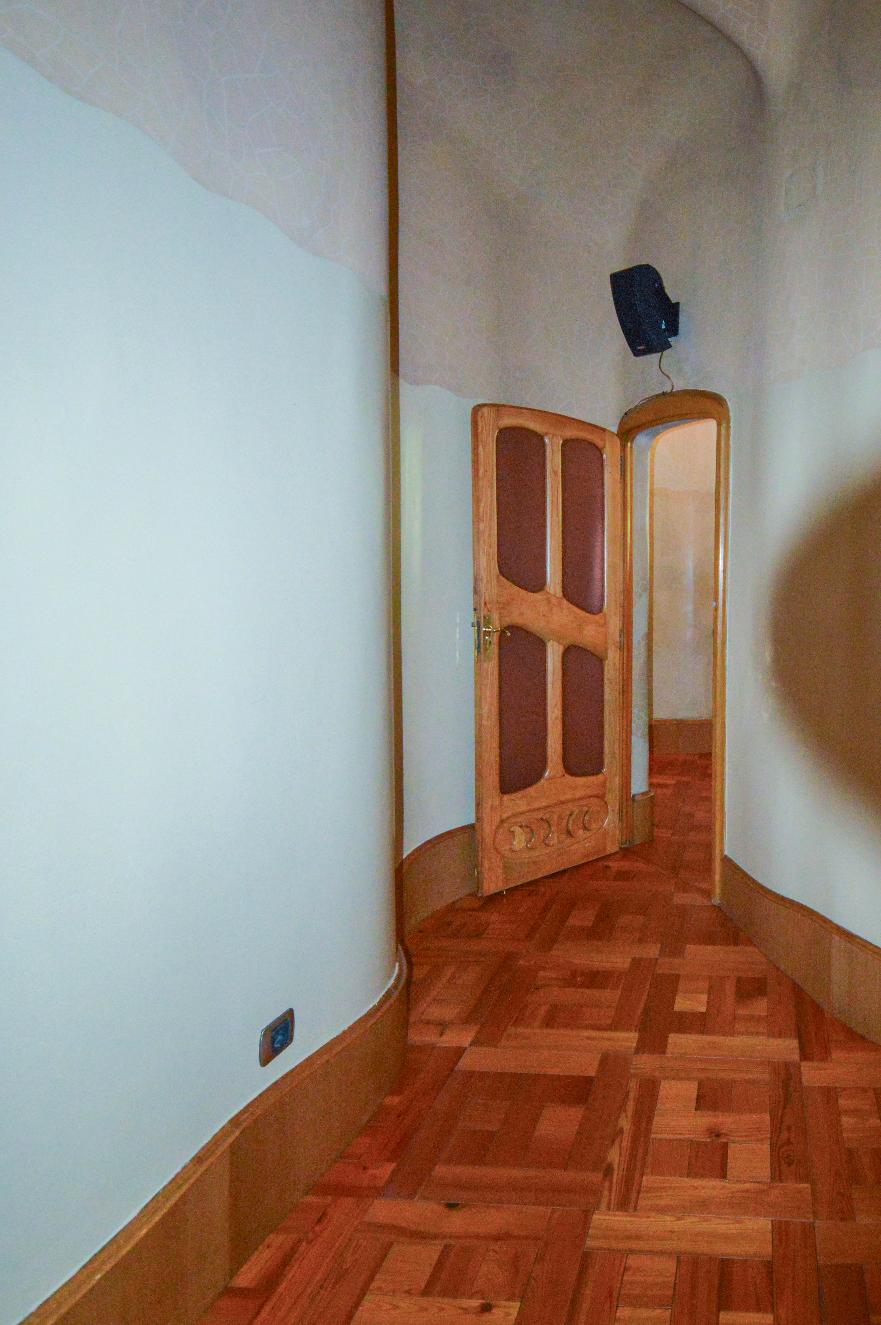 The image depicts a narrow hallway with a curved ceiling and wooden parquet flooring. The hallway leads to a wooden door with intricate designs and glass panels. The walls are painted white, and there is a light fixture mounted on the ceiling near the door.
